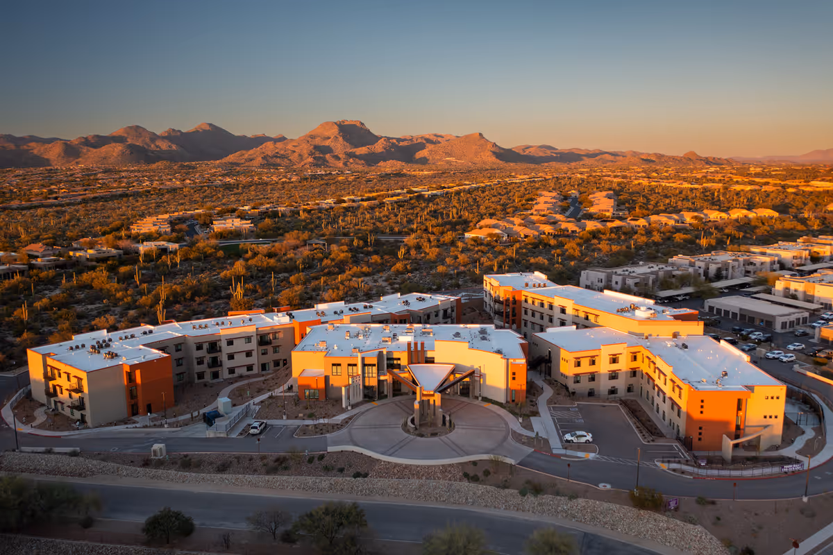 Aerial view of La Sonora at Dove Mountain senior living facility at sunset, showing a large, modern multi-wing building surrounded by desert landscape and mountains in the background.