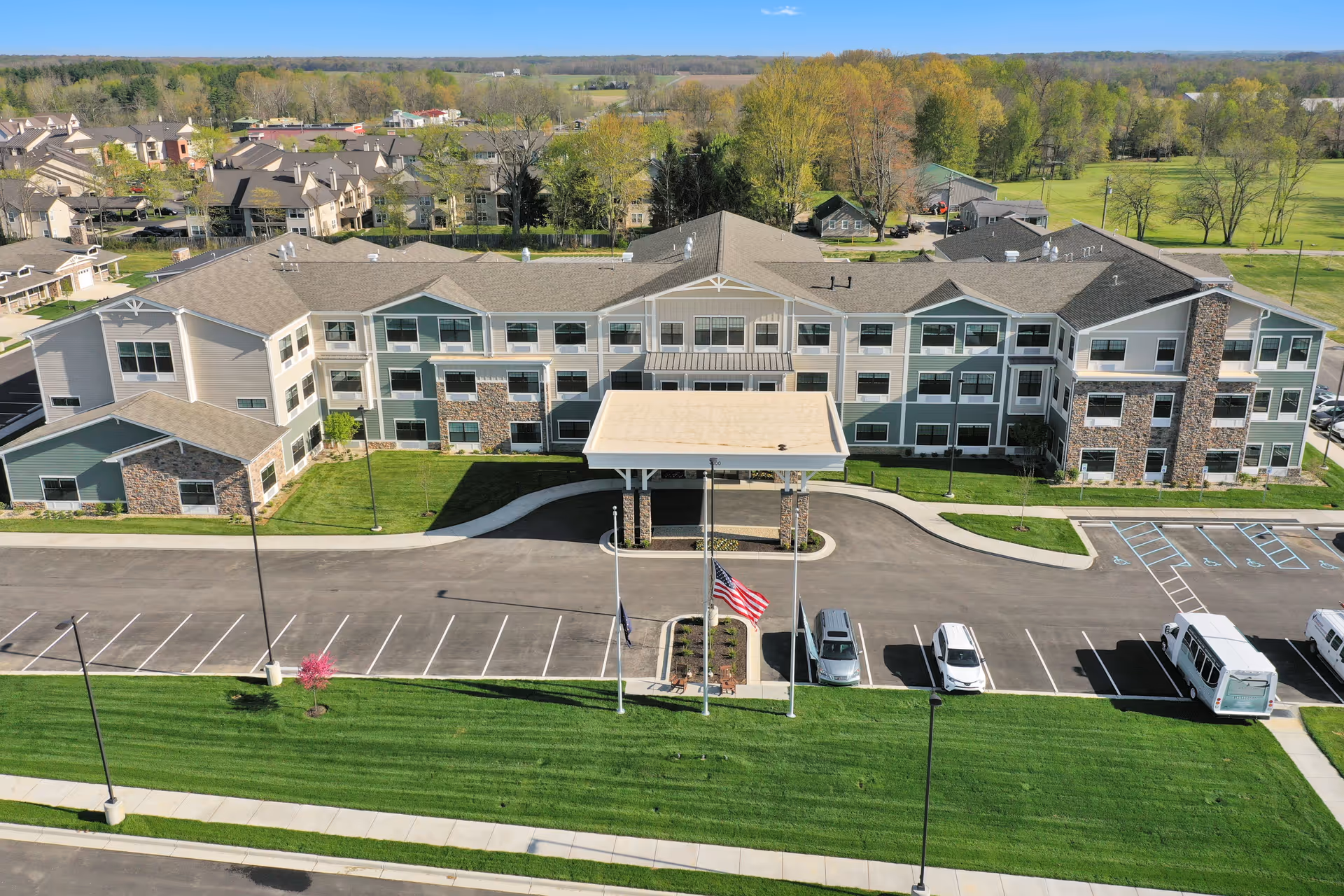 Aerial view of a three-story senior living building with a covered main entrance, adjacent parking lot, flagpoles, and green lawn.