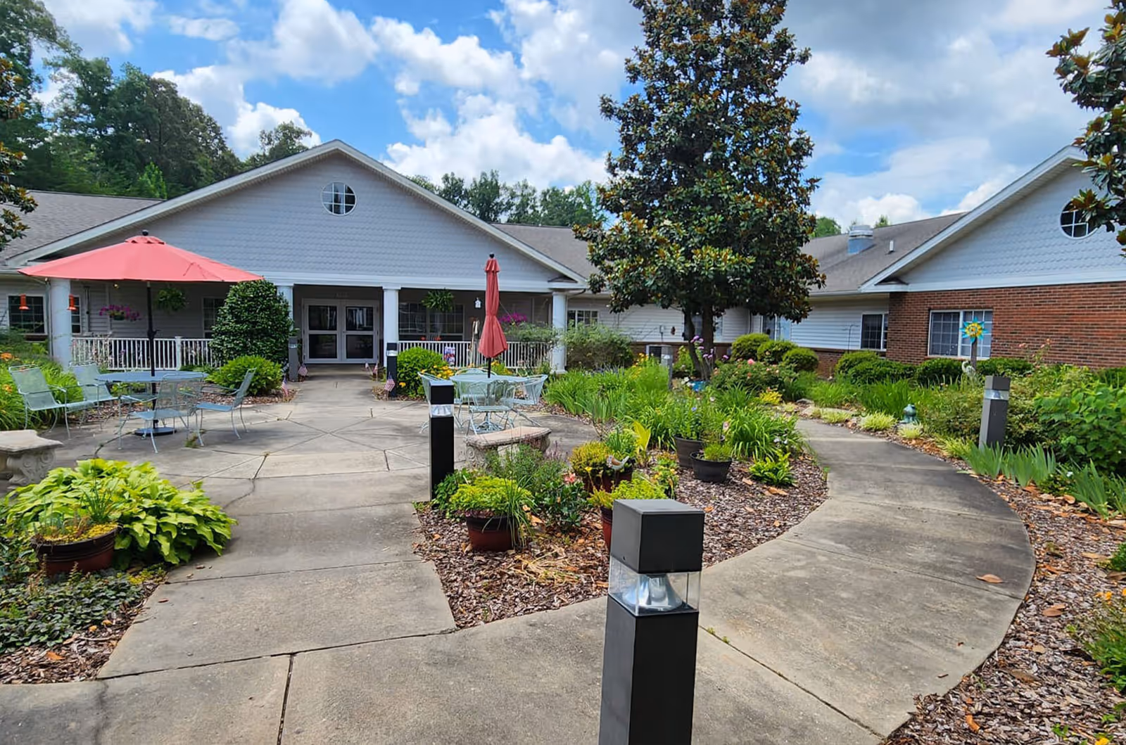 Outdoor garden area with a concrete pathway leading to the entrance of a single-story building. The garden features various plants, shrubs, and small trees, with patio tables and chairs under red umbrellas on the left side. The building has white siding with a brick section on the right and a covered porch at the entrance.