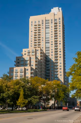 Tall beige residential building with multiple balconies under a clear blue sky, surrounded by green trees and a street with cars in the foreground.