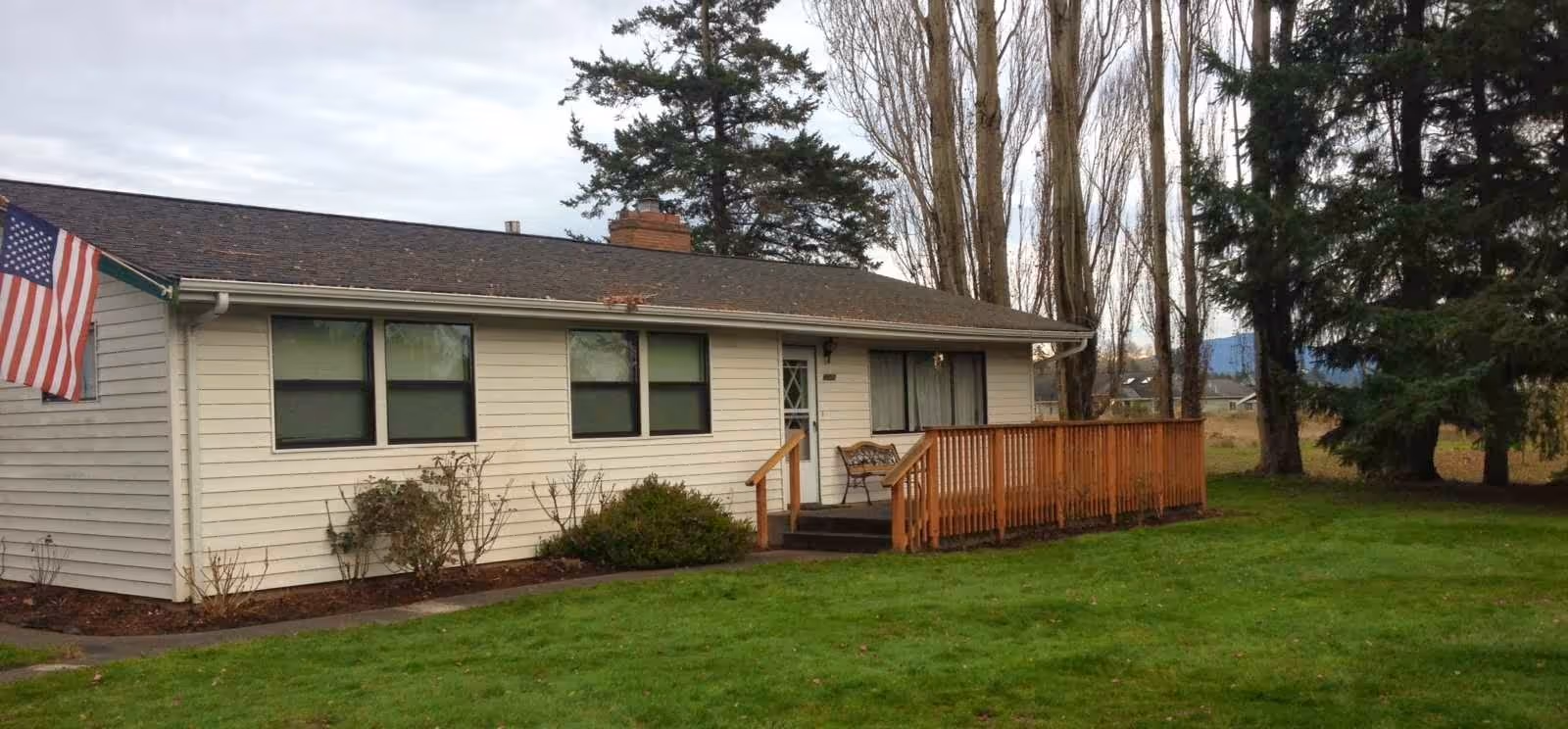 Single-story house with beige siding, a small wooden porch with a bench, several windows, an American flag mounted near the roof, and a large grassy yard with trees in the background under a cloudy sky.