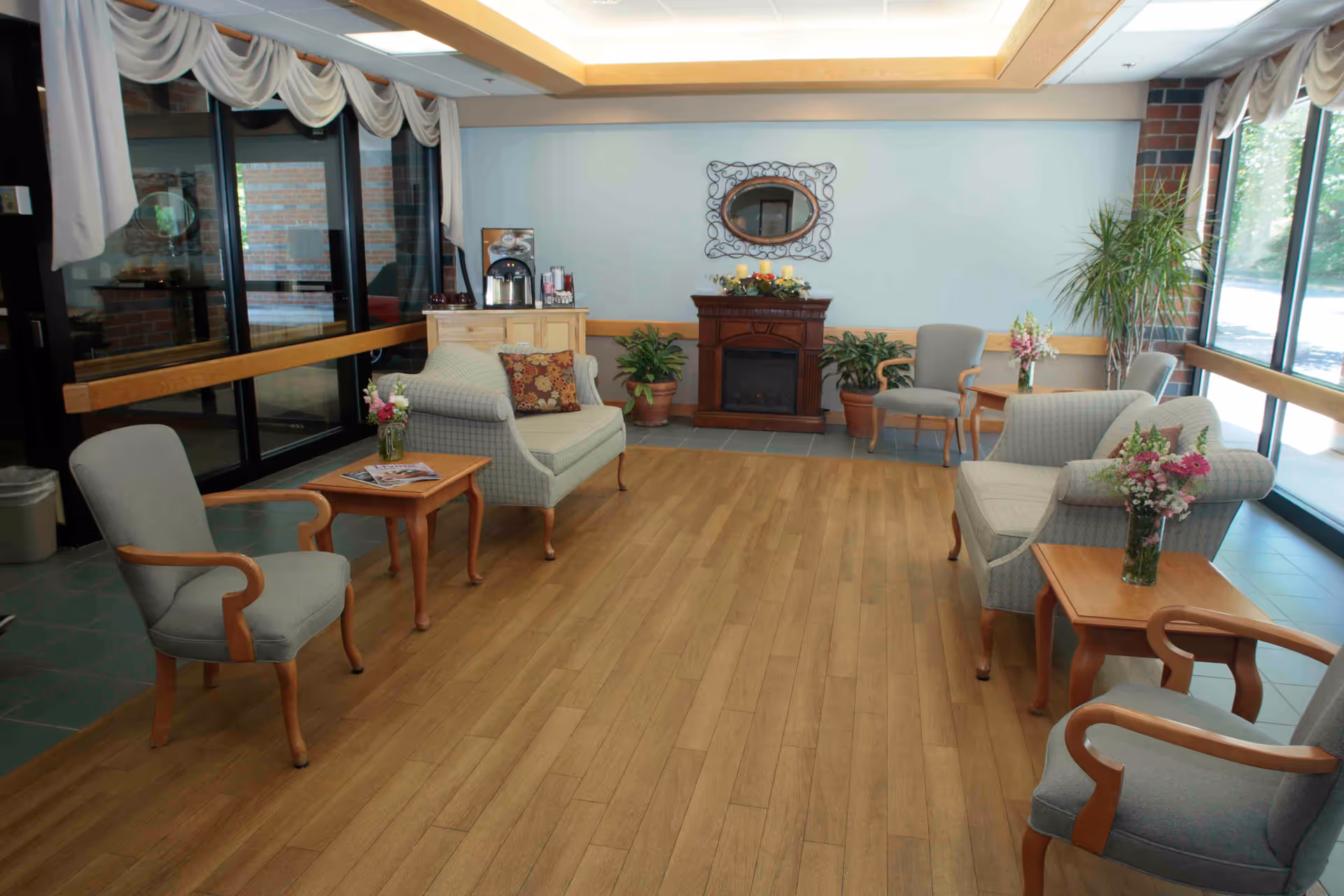 A cozy seating area in a senior living facility with several upholstered chairs and loveseats arranged around wooden side tables holding flower vases. There is a small electric fireplace against the far wall with a decorative mirror above it and potted plants on either side. Large windows let in natural light, and the floor is a combination of wood and tile.