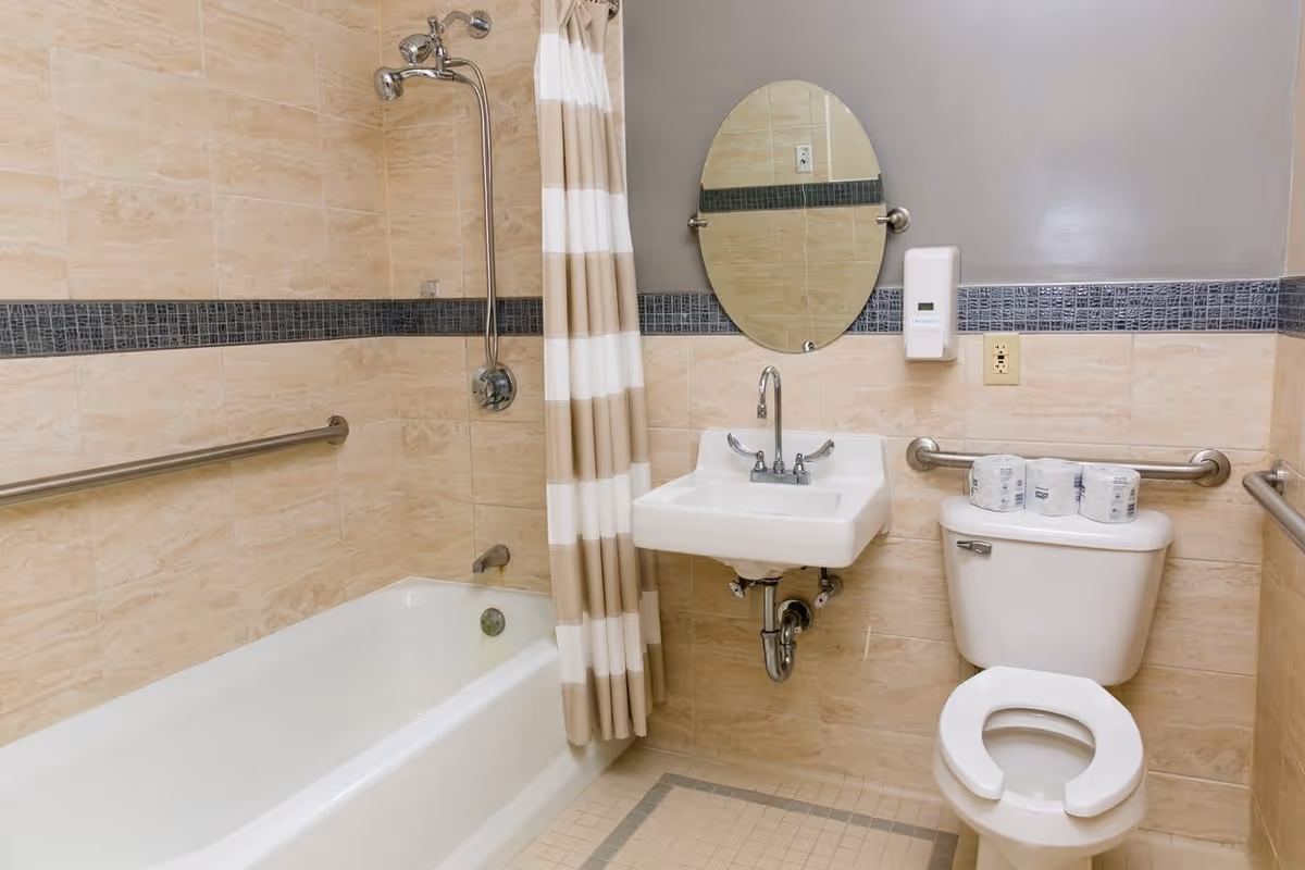 A clean bathroom with beige tiled walls and floor, featuring a white bathtub with a striped shower curtain, a wall-mounted sink with an oval mirror above it, a soap dispenser, and a toilet with three rolls of toilet paper on the tank. There are metal grab bars installed near the bathtub and toilet for accessibility.
