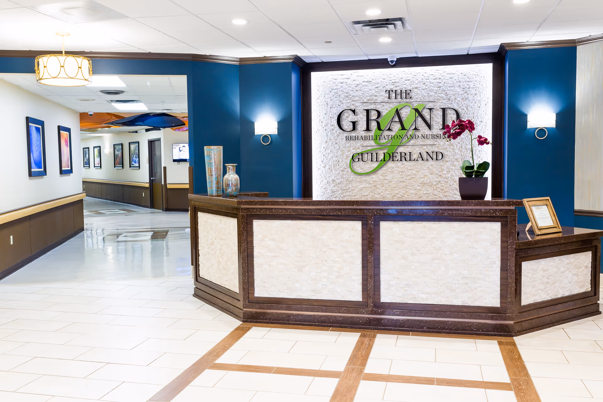 Reception area of The Grand Rehabilitation and Nursing at Guilderland featuring a front desk with decorative vases and a potted orchid, a textured wall with the facility's name, blue accent walls, and a hallway with framed artwork.