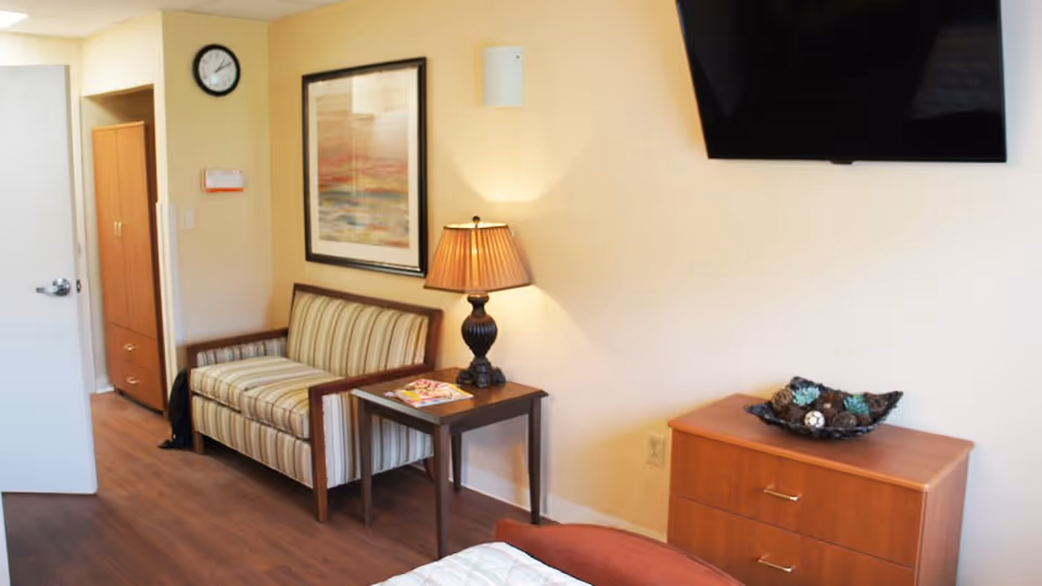 A furnished nursing facility bedroom with a striped loveseat, side table and lamp, wall-mounted TV, and a wooden dresser.