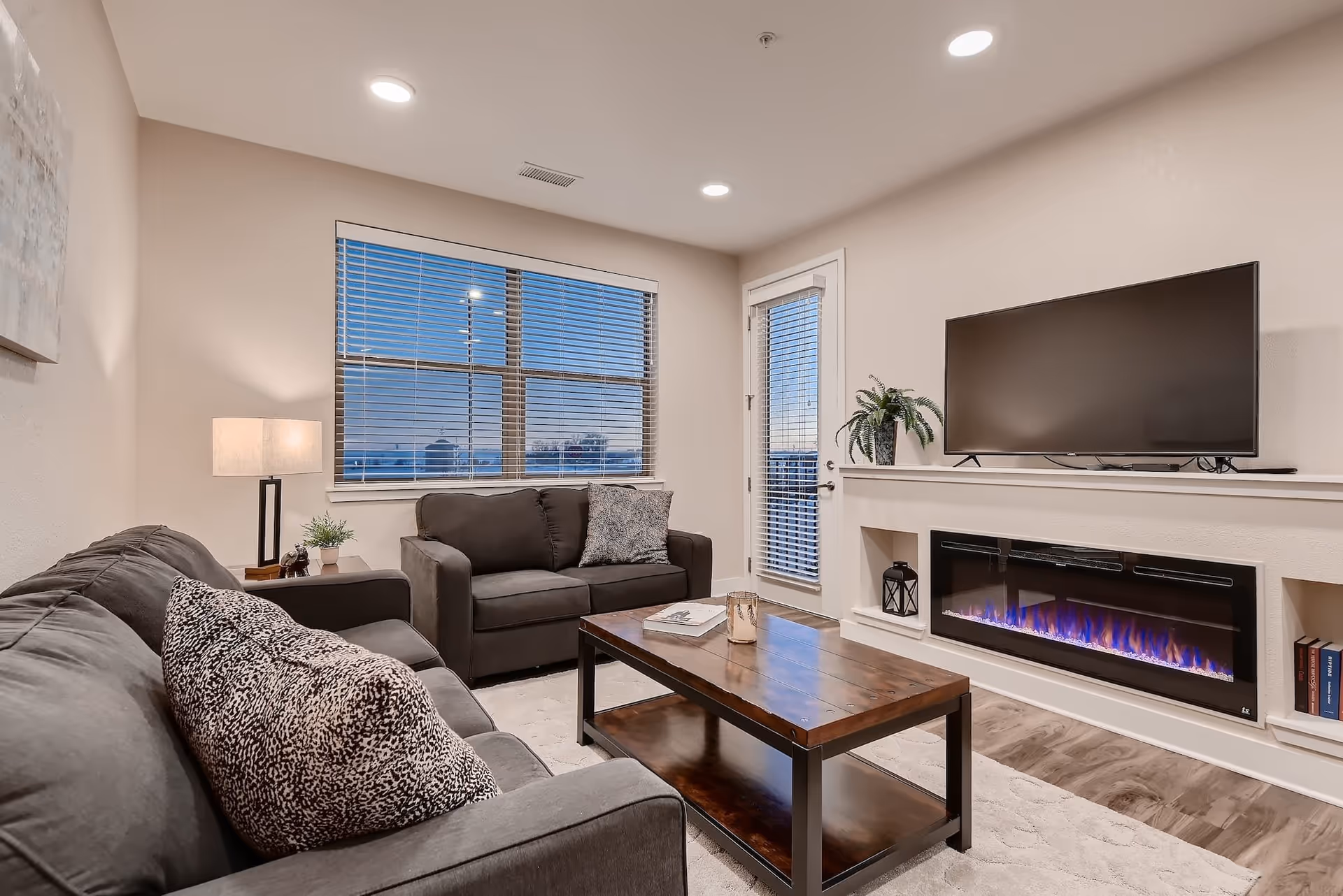 A cozy living room with two dark gray sofas, a wooden coffee table, a modern electric fireplace with blue flames, a flat-screen TV mounted above the fireplace, a table lamp, and a window with blinds showing an evening sky.