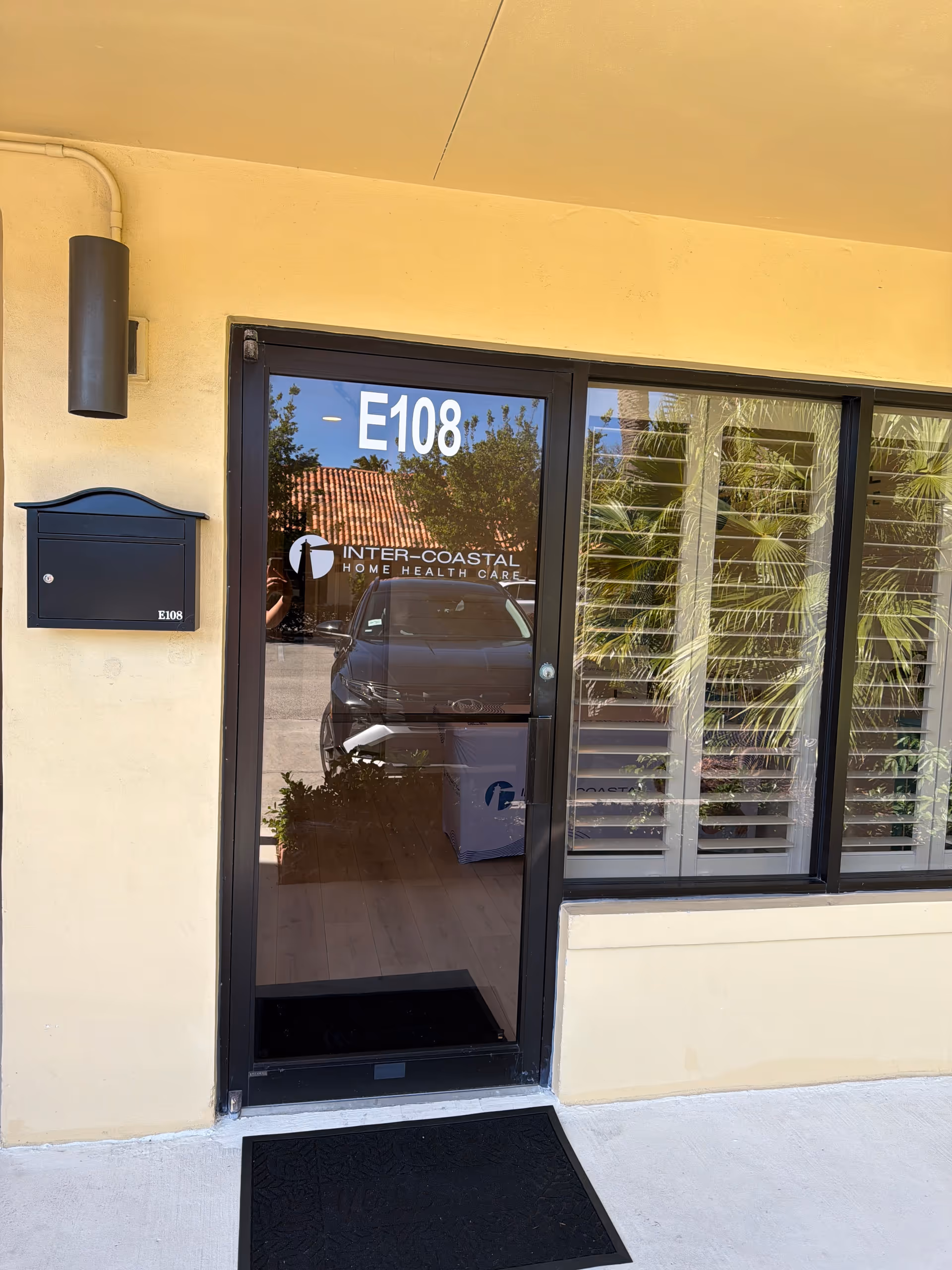 Clear glass door entrance labeled E108 for Inter-Coastal Home Health Care, with reflected car and plants.