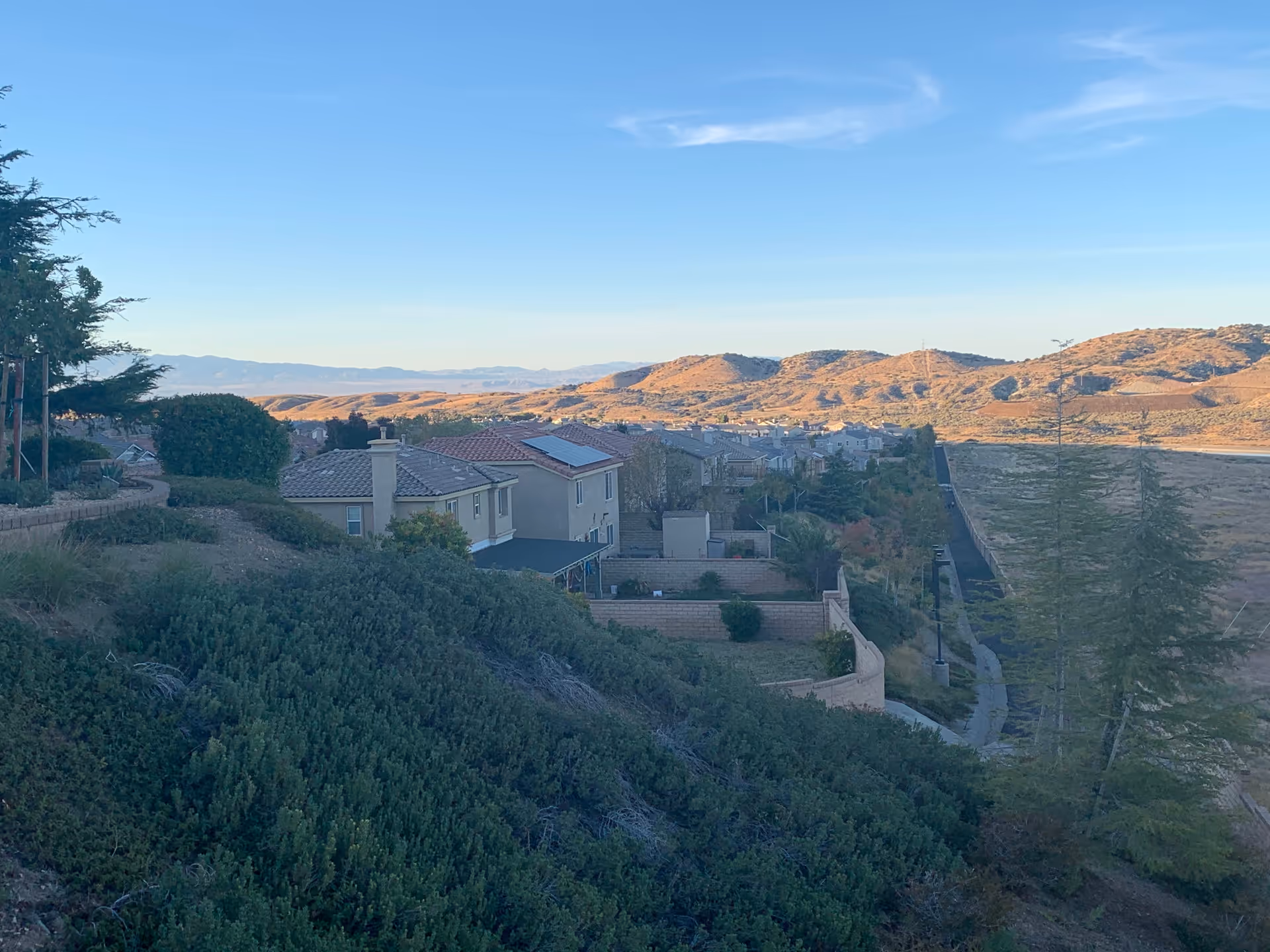 View of a residential area with houses featuring tiled roofs, surrounded by greenery and hills in the background under a clear blue sky.