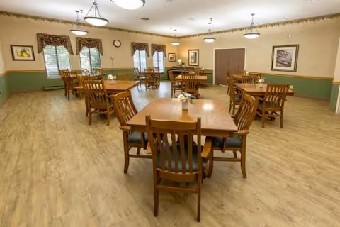 A spacious dining room with multiple wooden tables and chairs arranged neatly. The room has wood flooring, green and beige walls, framed artwork, and windows with valances. Ceiling lights provide illumination.