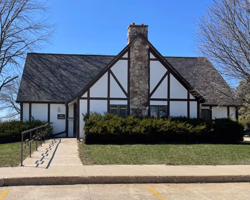 Tudor-style building with white walls, dark trim, central stone chimney, and trimmed hedges.