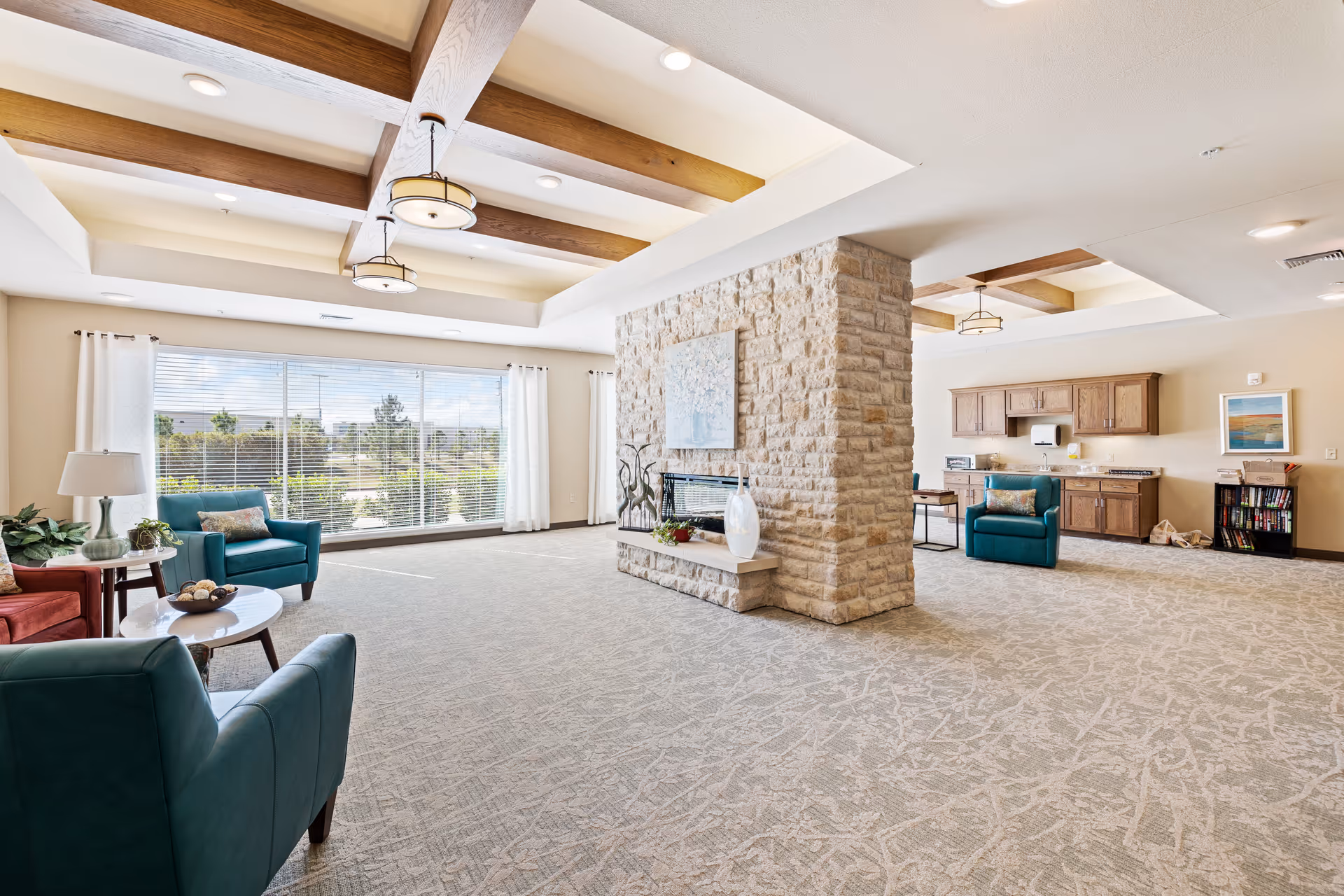 Spacious senior living common area with teal chairs, stone column, large windows, and kitchenette.