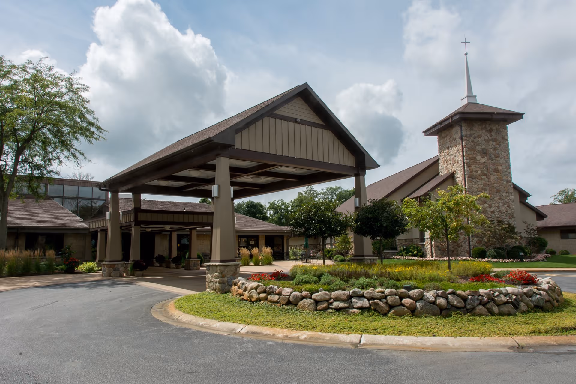 Exterior view of a senior living facility with a covered entrance supported by stone pillars, a circular driveway, landscaped garden with rocks and flowers, and a building with a stone tower topped by a cross under a partly cloudy sky.