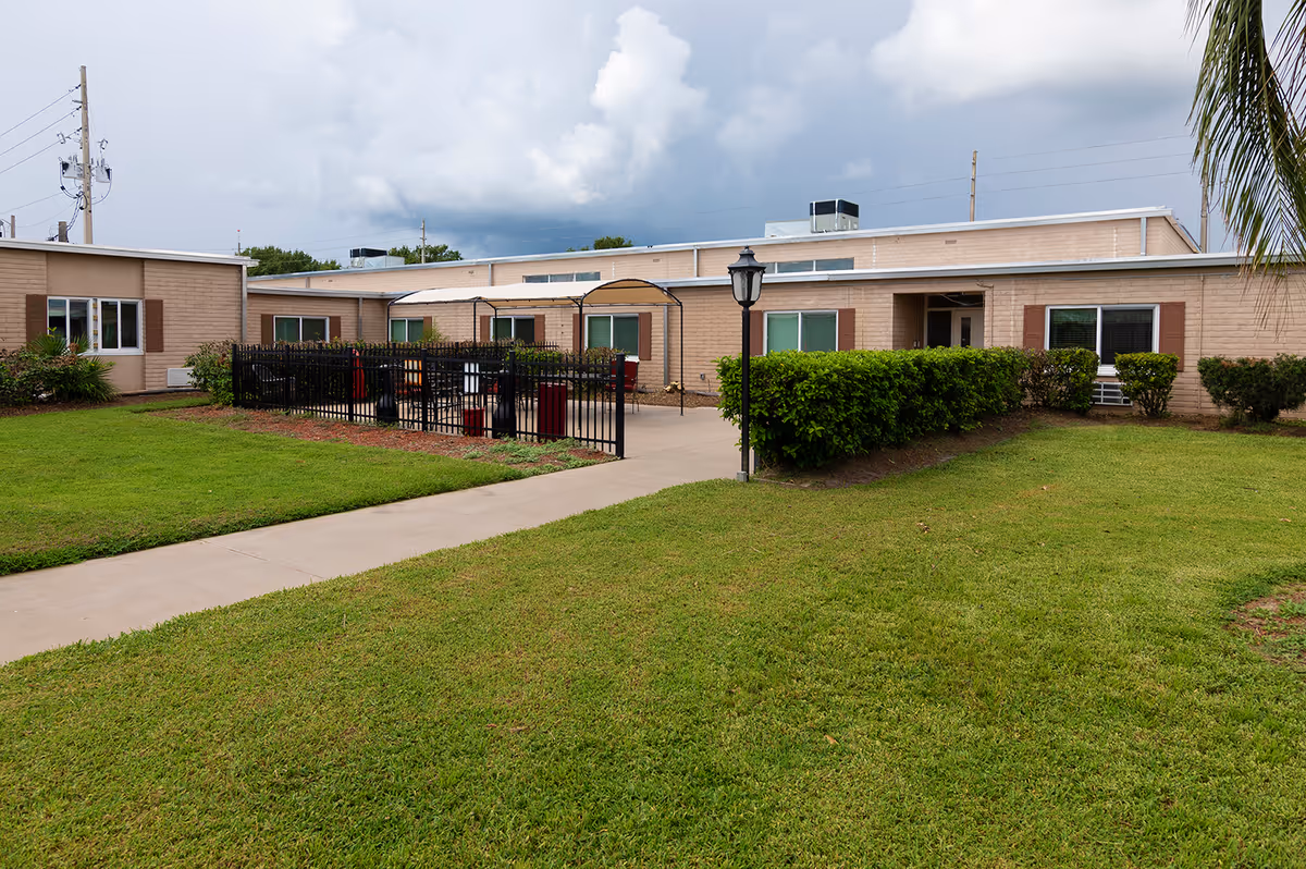 Exterior front of a single-story senior living building with a fenced courtyard, walkway, lamp post, and lawn under a cloudy sky.