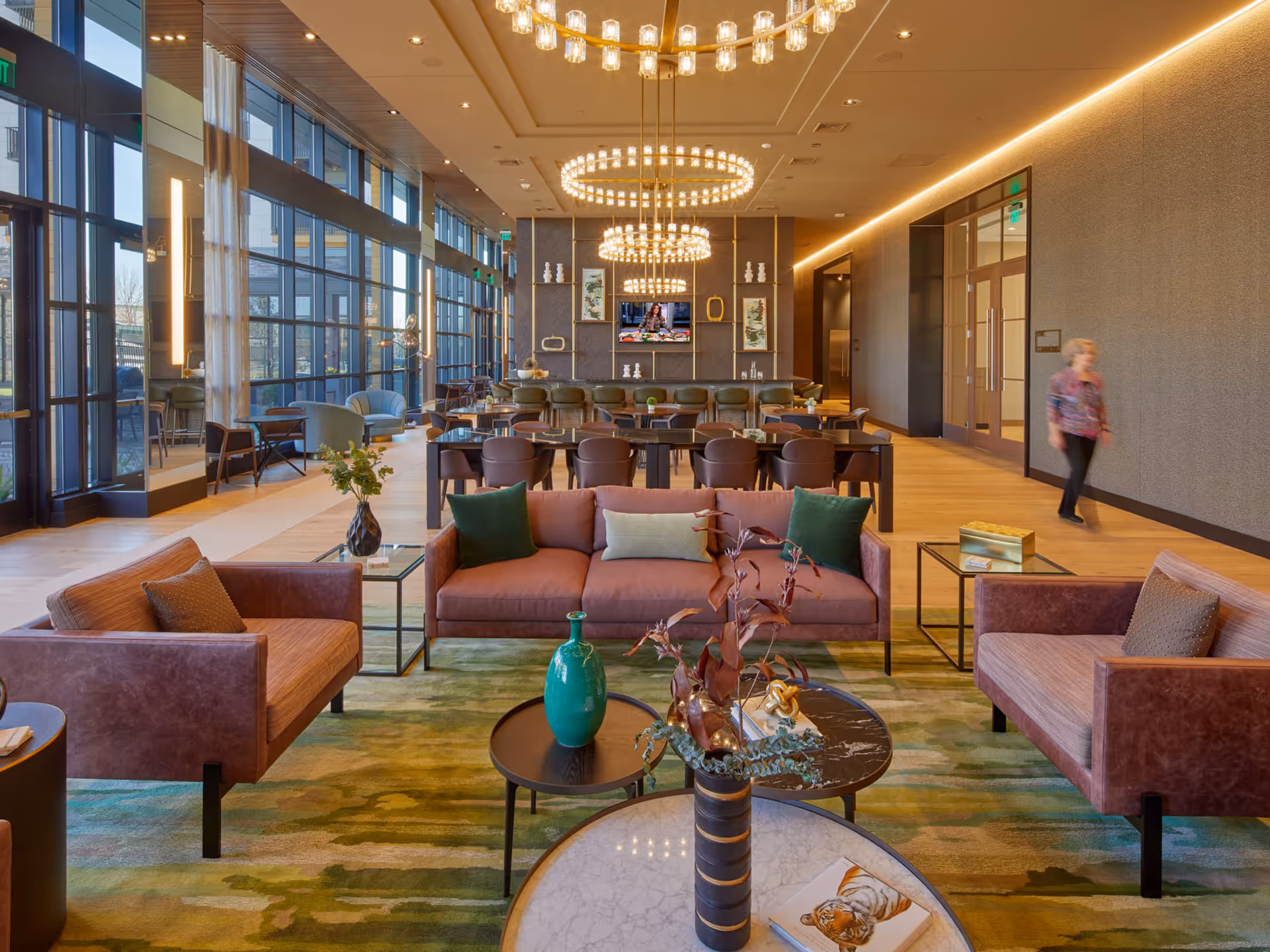 A spacious and modern common area in a senior living facility featuring comfortable seating with two armchairs and a sofa around a set of coffee tables with decorative vases and a book. Behind the seating area is a large dining table with multiple chairs and a long counter with bar stools. The room has large floor-to-ceiling windows on the left side allowing natural light to fill the space. Three circular chandeliers hang from the ceiling, and a woman is walking along the right side of the room.