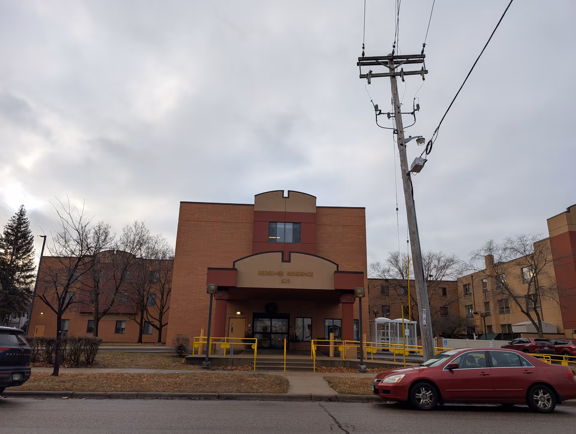 Front exterior of a brick senior residence building with an entrance canopy, wheelchair ramp, and parked cars under an overcast sky.