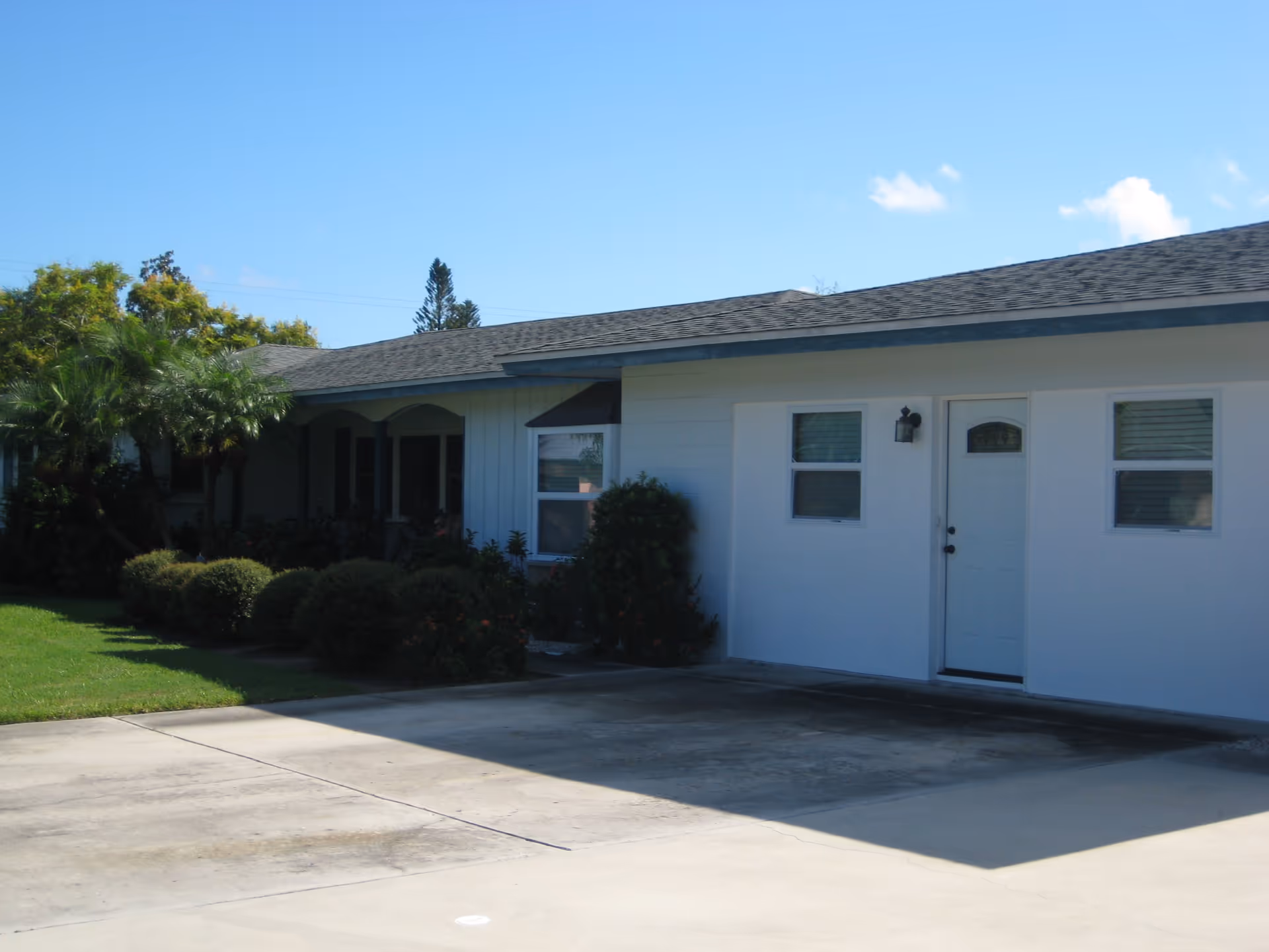 Single-story white building front with a driveway, front door, windows and landscaped shrubs under a clear blue sky.