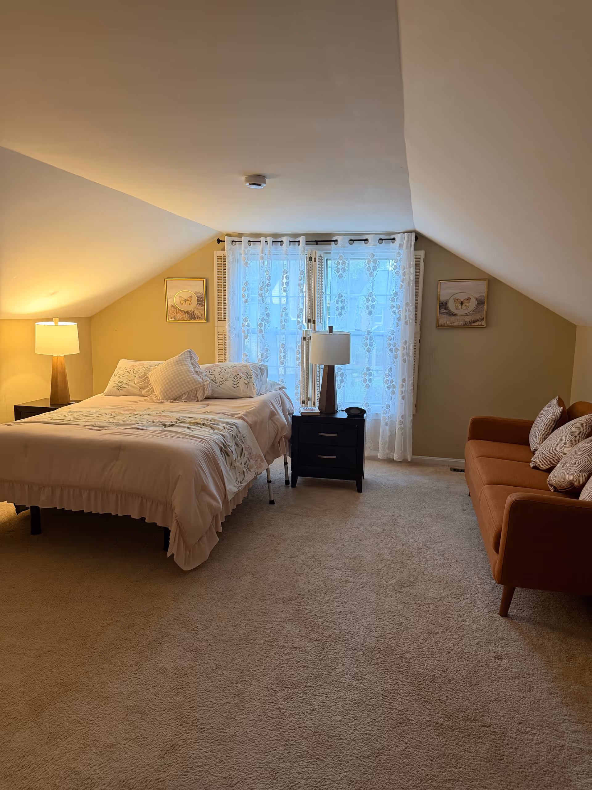 Bedroom with bed, nightstands, lamps, windowed wall, and a sofa under a sloped ceiling.