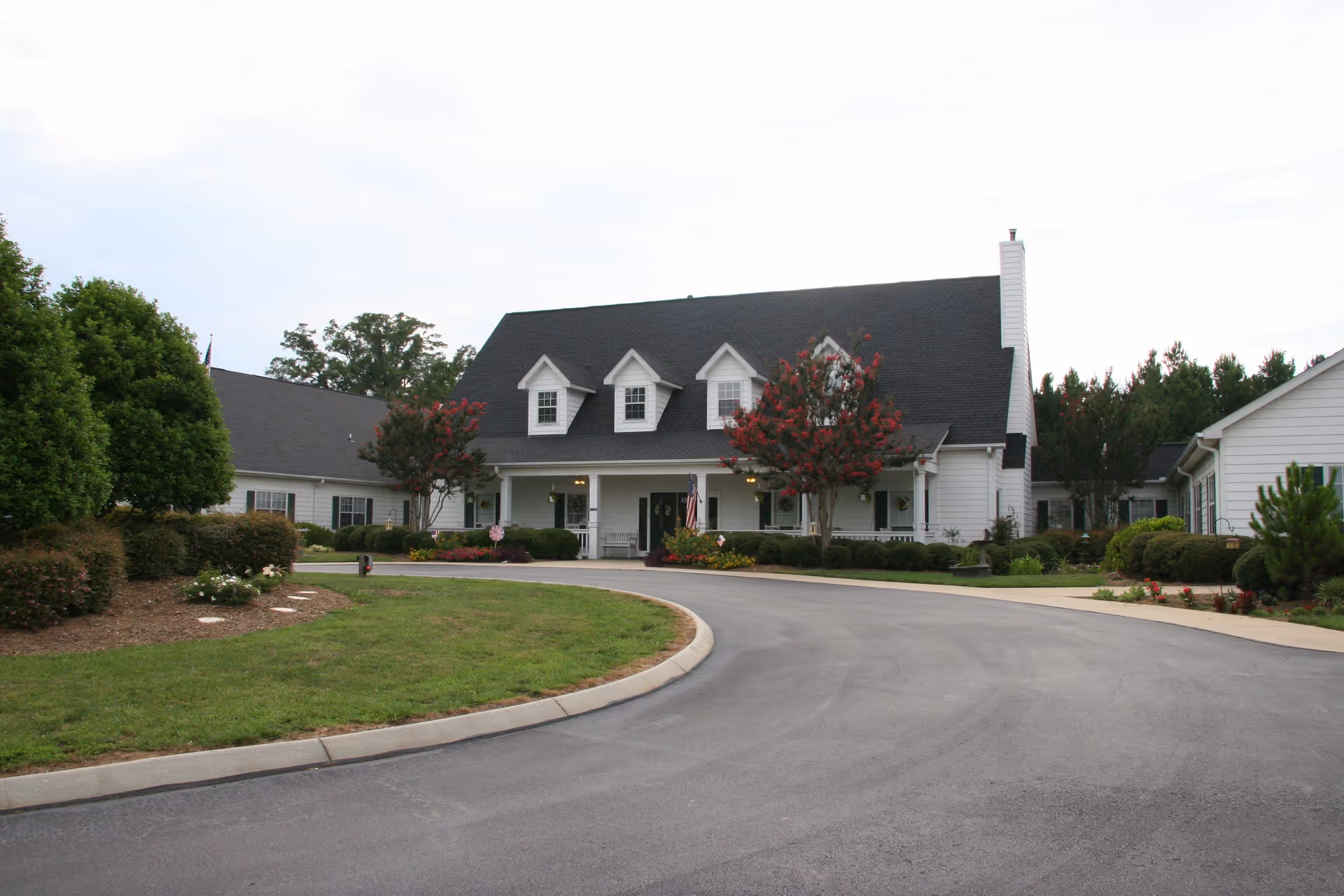 Front exterior view of a large white building with a black roof, featuring three dormer windows and a covered porch with seating. The building is surrounded by well-maintained landscaping, including bushes, trees with red flowers, and a curved driveway leading up to the entrance.