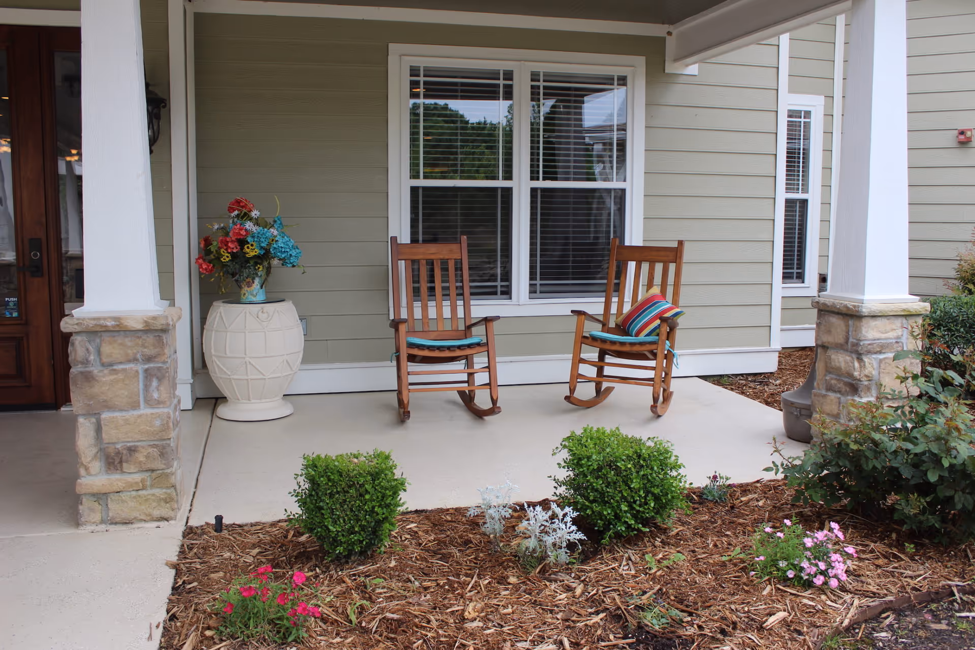 Covered front porch with two wooden rocking chairs, a large planter of flowers, and small garden beds in front of a window.