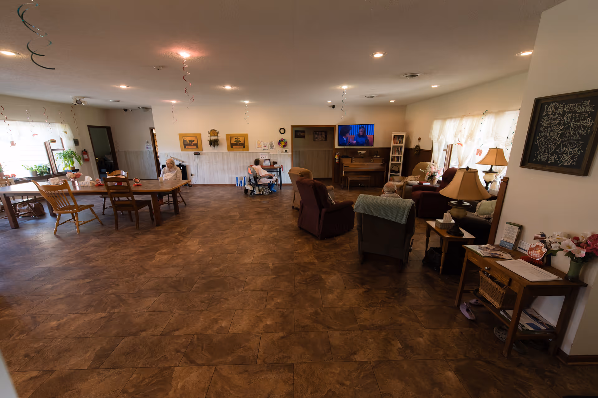 A spacious common area in a senior living facility with several elderly residents seated in chairs and wheelchairs. The room features a wooden dining table with chairs on the left, a television mounted on the wall, a piano, and comfortable armchairs arranged near windows with sheer curtains. The floor is covered with brown tiles, and the walls are decorated with framed pictures and a chalkboard with writing. There are also some lamps and flower arrangements on side tables.