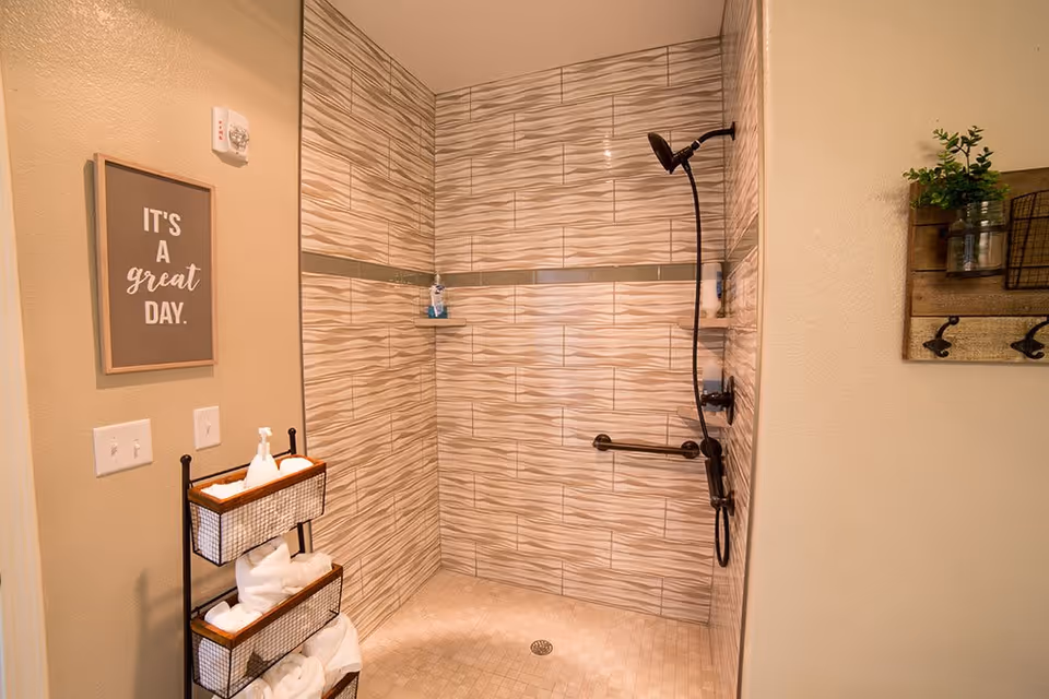 A walk-in shower with beige and brown wave-patterned tiles on the walls and a beige tiled floor. The shower has a black showerhead and a grab bar. On the left wall outside the shower, there is a framed sign that reads 'IT'S A great DAY.' Below the sign is a black metal rack holding white towels and a soap dispenser. On the right wall, there is a wooden wall-mounted shelf with hooks and a small plant in a glass jar.