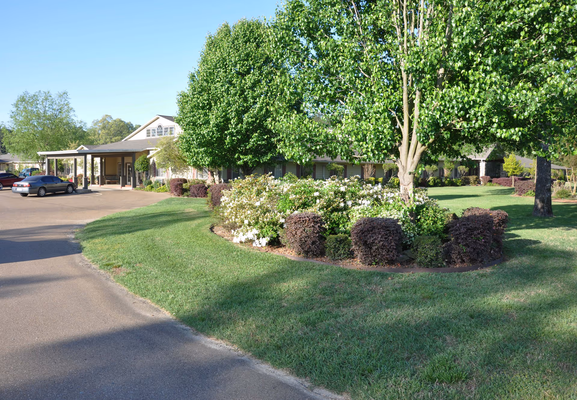 Exterior view of Dudneywood Assisted Living facility showing a driveway, well-maintained lawn, landscaped garden with bushes and trees, and the building entrance with a covered porch.