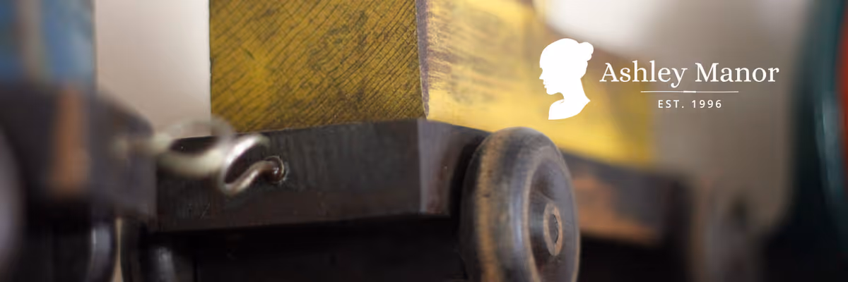 Close-up image of a wooden cart or piece of furniture with a yellow painted section and a small wheel, with the Ashley Manor logo and text 'Ashley Manor EST. 1996' in the upper right corner.