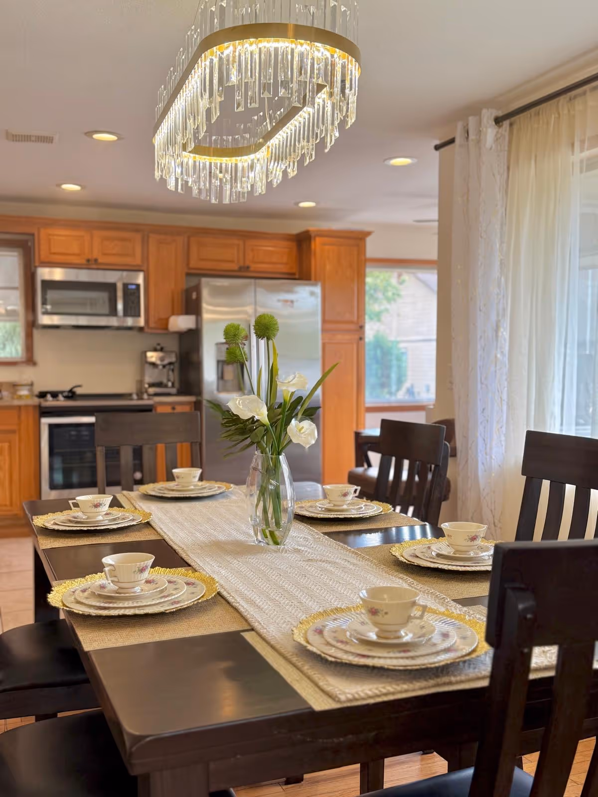 Wood dining table set for six under a crystal chandelier in a bright kitchen dining area.