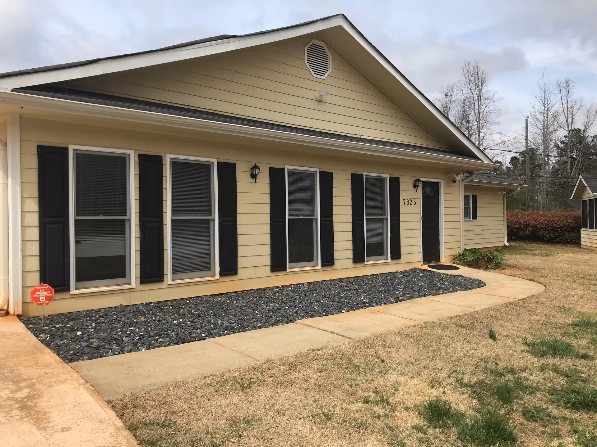 Front exterior of a single-story yellow house with multiple windows with black shutters and a concrete walkway leading to the entrance.