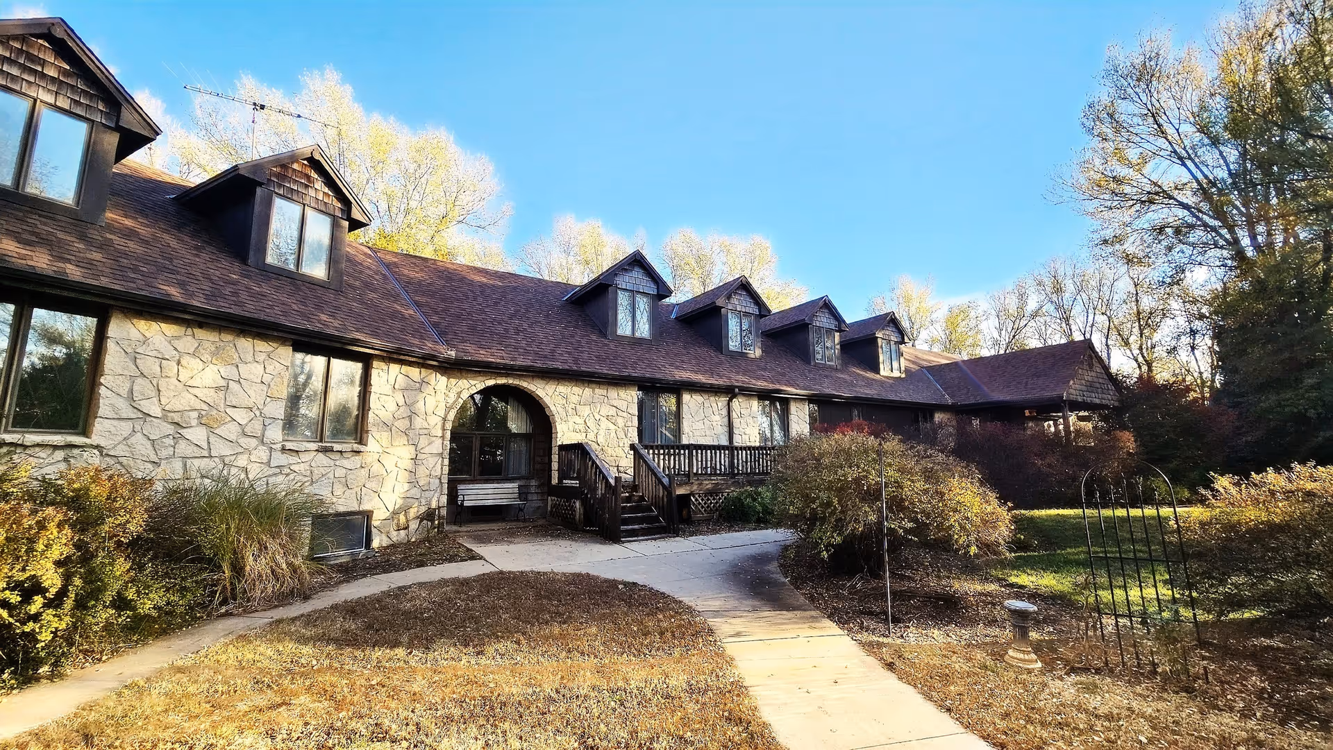 Stone-faced two-story building with dormer windows, a front porch, and a curved walkway through landscaped grounds under a clear blue sky.