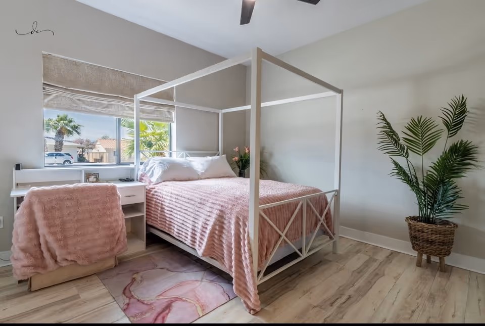 A pastel pink four-poster bedroom with a bed, desk, window, and potted plant.
