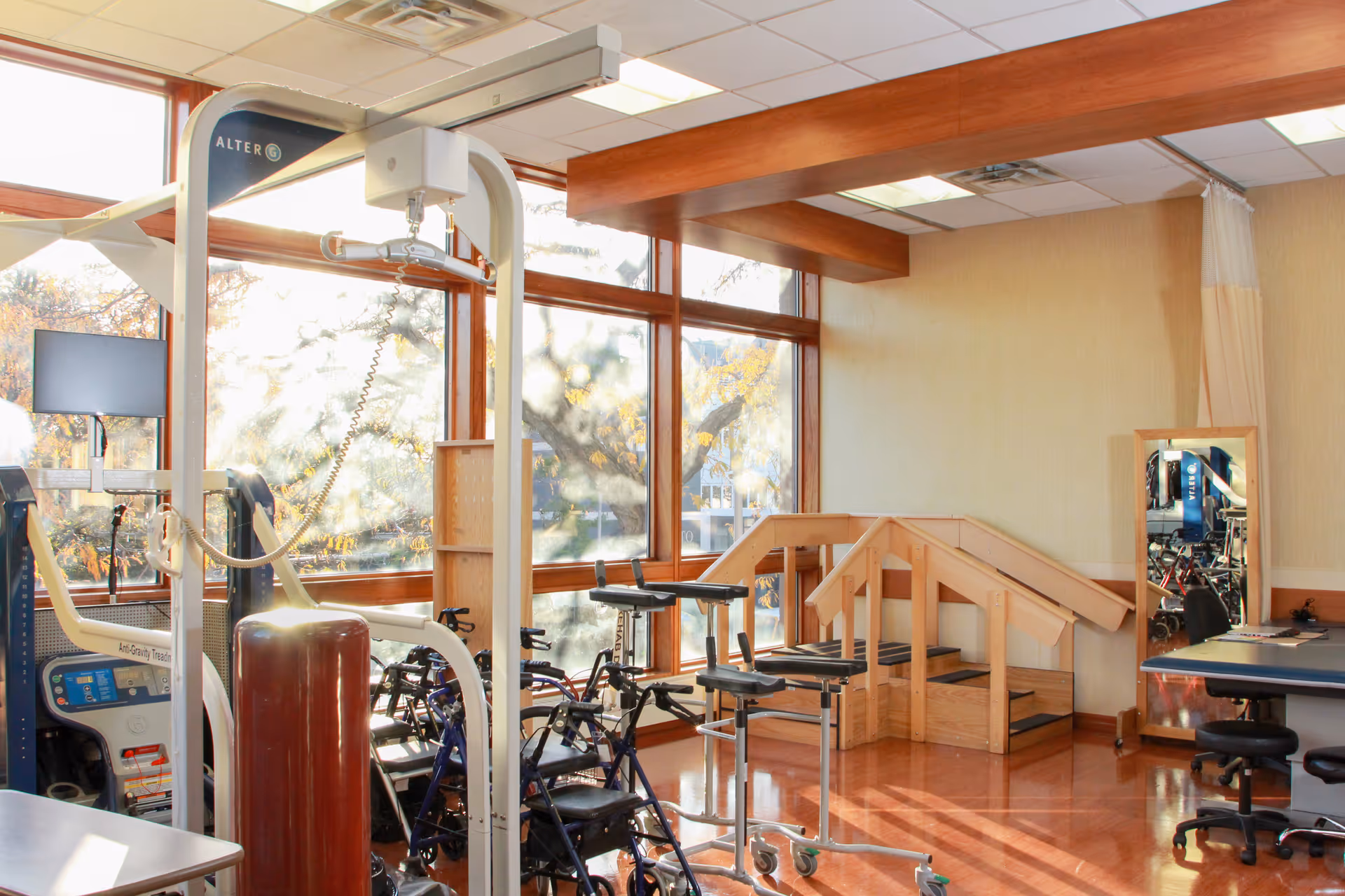 Bright rehabilitation gym with exercise machines, bikes, and a wooden therapy ramp near large windows.