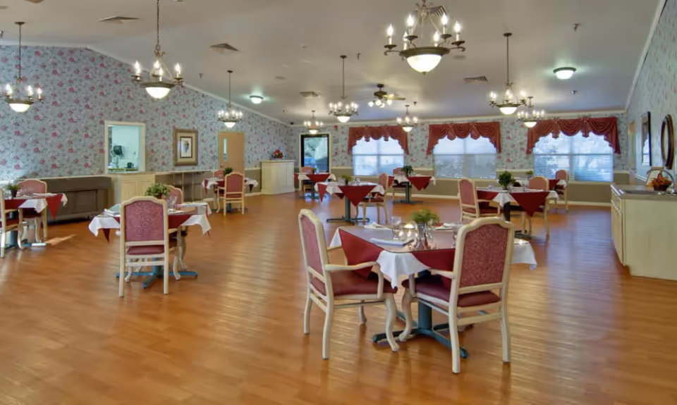 Spacious dining room with multiple set tables and chairs, chandeliers, floral wallpaper, and wood flooring.