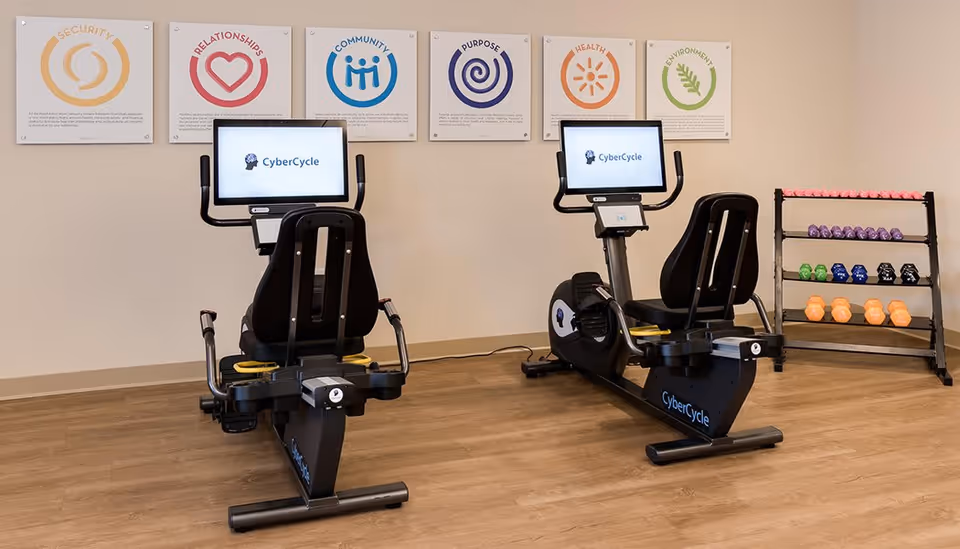 A fitness room with two CyberCycle recumbent exercise bikes facing a wall with six colorful posters displaying words and icons related to security, relationships, community, purpose, health, and environment. To the right, there is a rack holding various colorful dumbbells. The room has wooden flooring and a neutral-colored wall.
