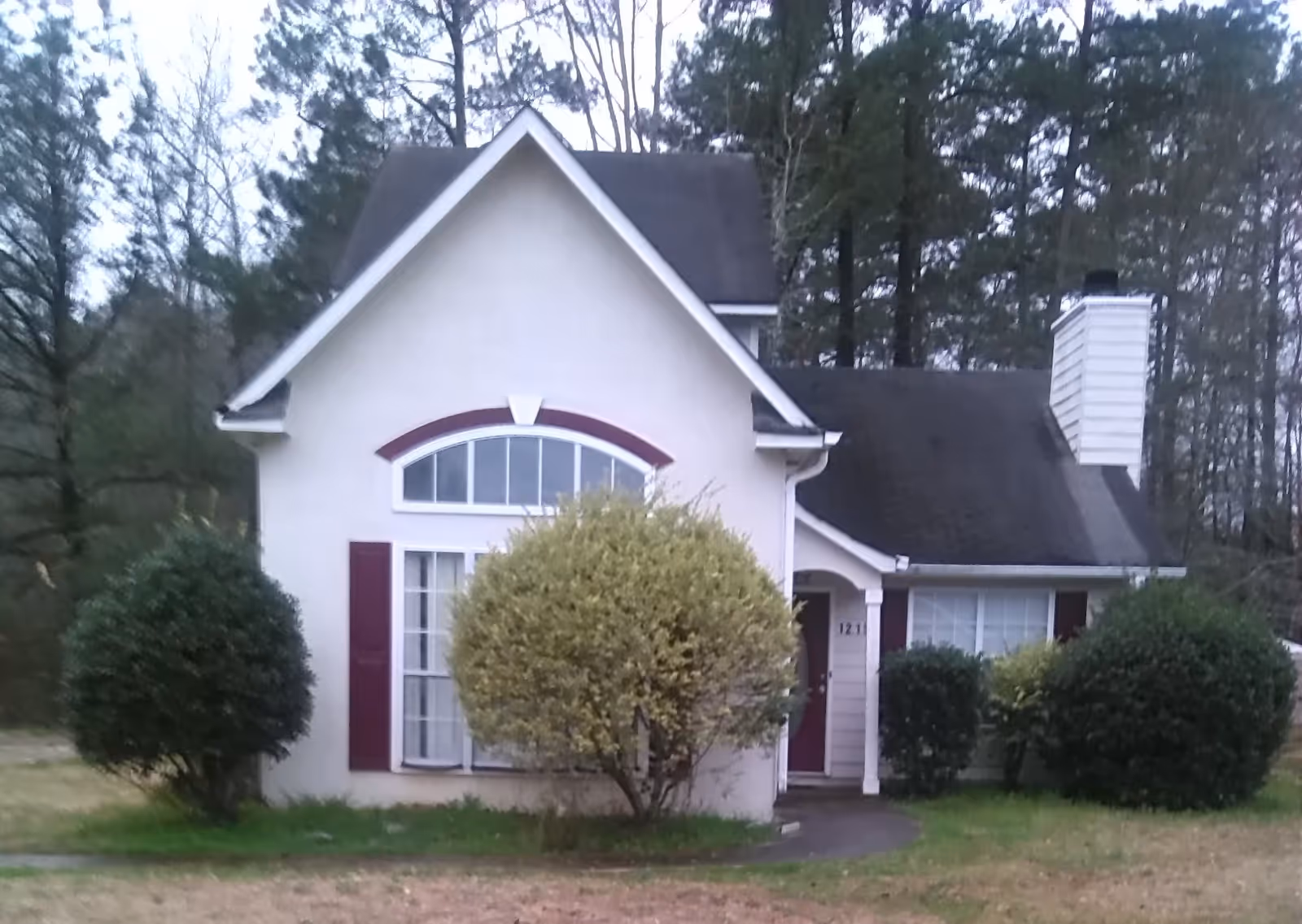 Front exterior view of a small house with a steep roof, a large arched window with red shutters, a front door partially visible, and several bushes in the front yard. Trees are visible in the background.