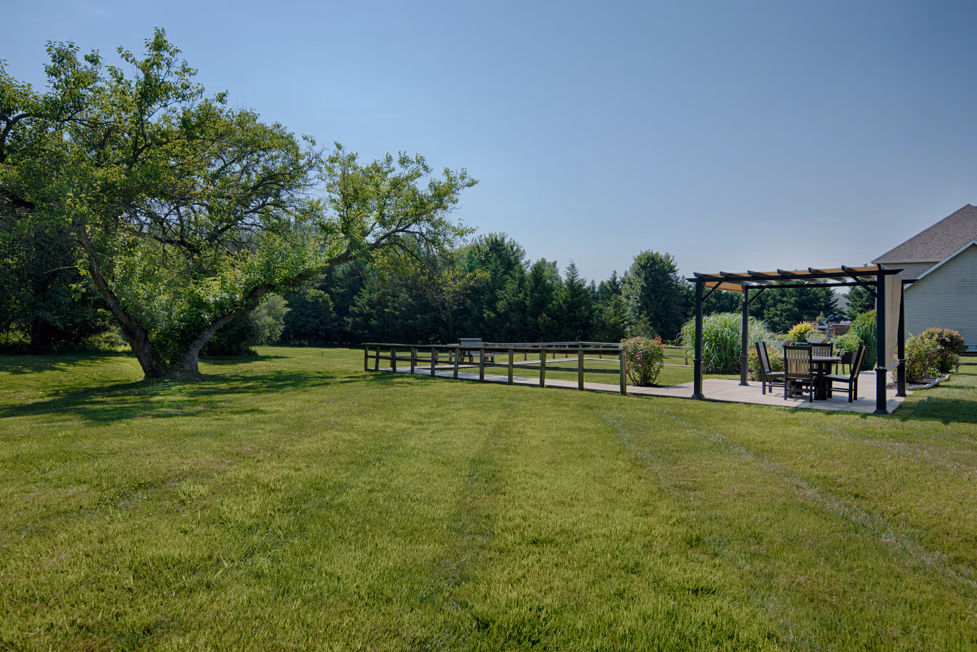 Spacious grassy lawn with a large tree, wooden fence, and a shaded outdoor dining pavilion.