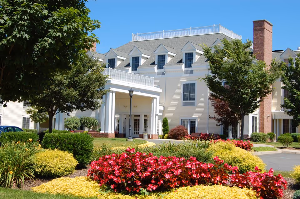 Front exterior of a large assisted living building with a columned entrance, manicured flower beds, and trees.