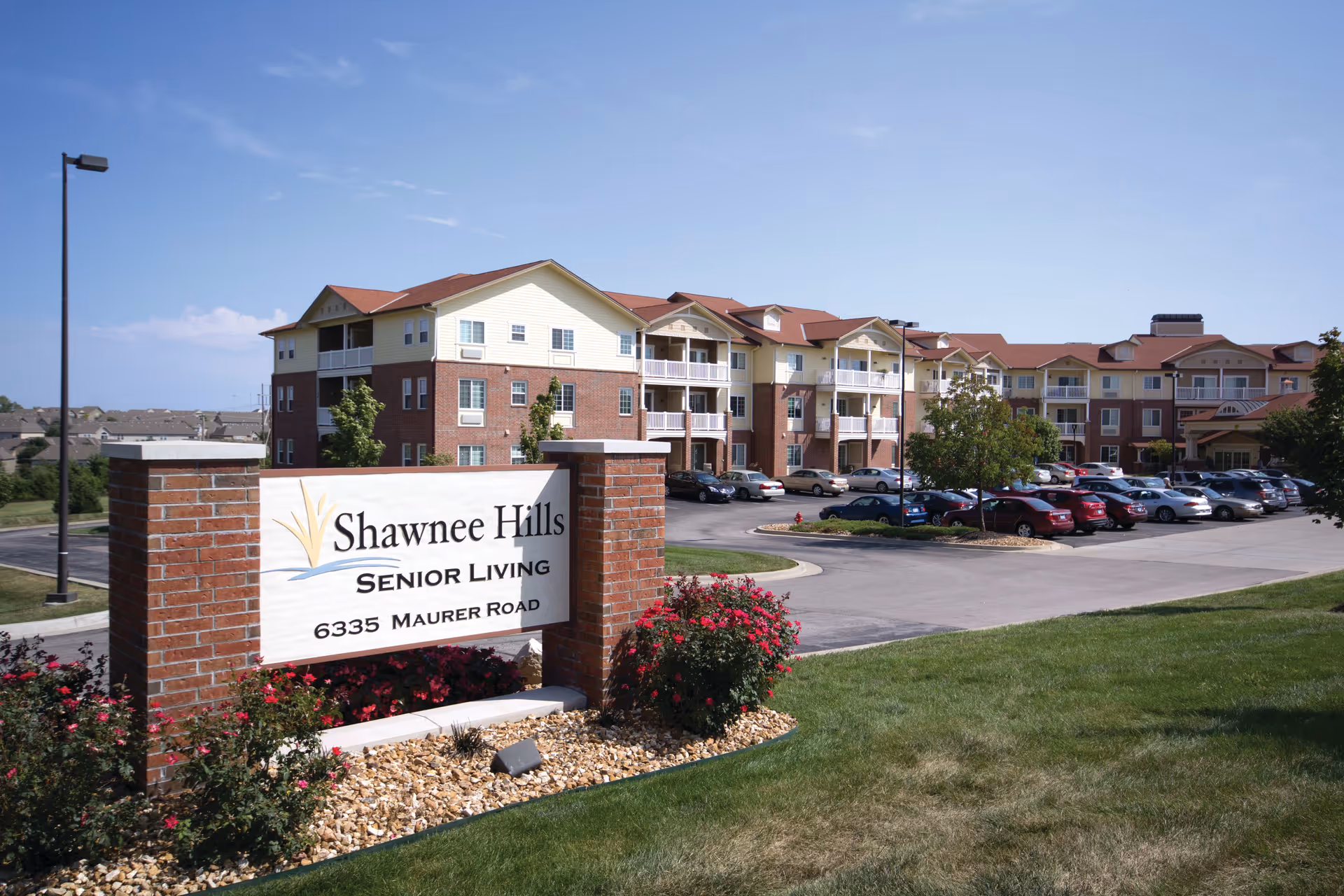 Exterior view of Shawnee Hills Senior Living facility showing a large multi-story building with balconies, a parking lot filled with cars, and a landscaped area with grass, bushes, and a sign displaying the facility's name and address.