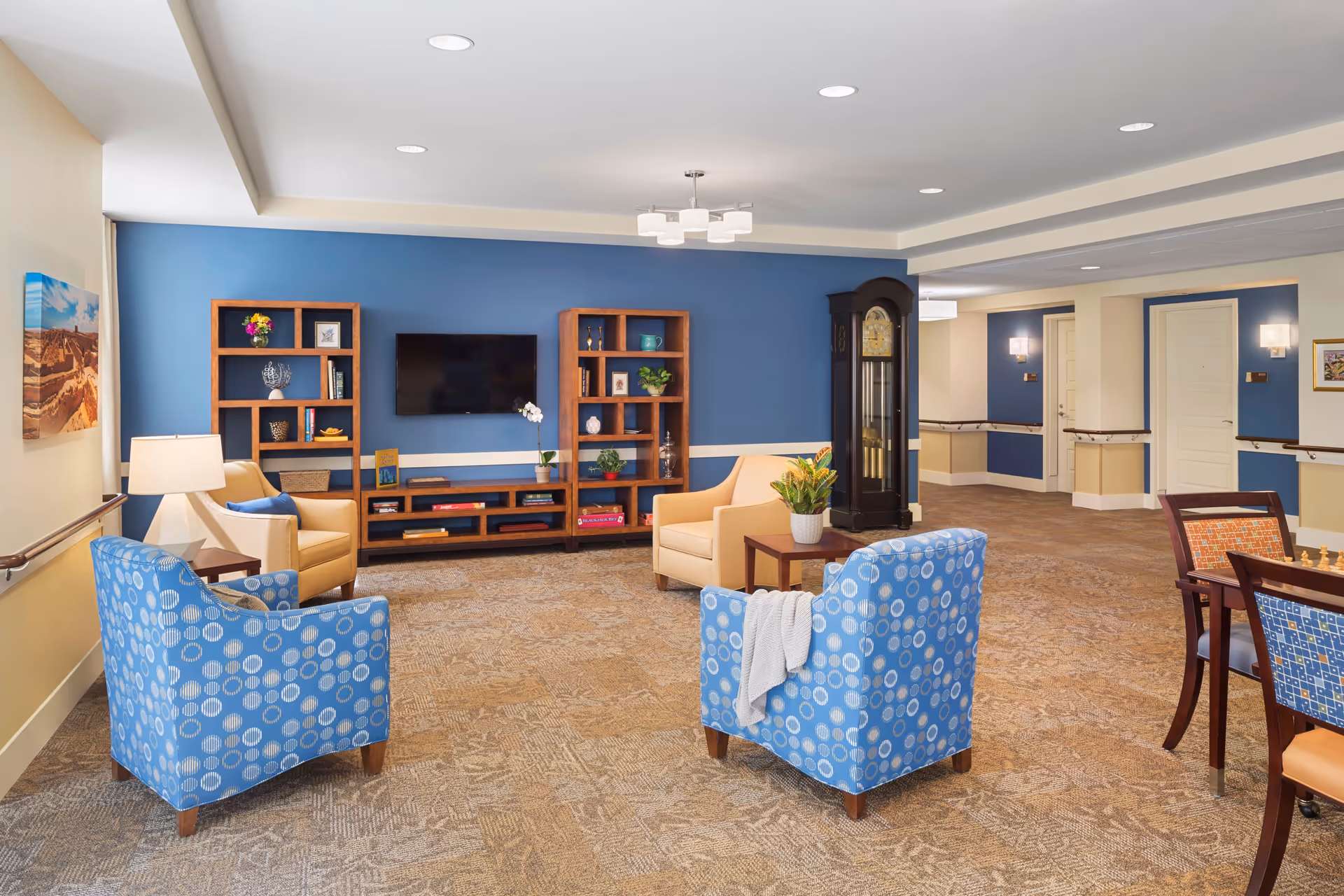 A cozy senior living common area with blue and beige walls, featuring two blue patterned armchairs and two beige armchairs arranged around a small wooden table with a plant. There are wooden shelves with decorative items and a flat-screen TV mounted on the blue wall. A grandfather clock stands near the hallway with white doors and wall sconces. A chessboard is visible on a table to the right.