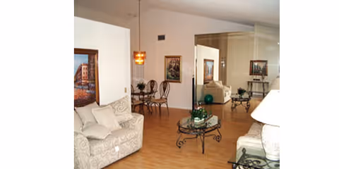A spacious living room in a senior facility with beige sofas, glass coffee tables, and artwork.