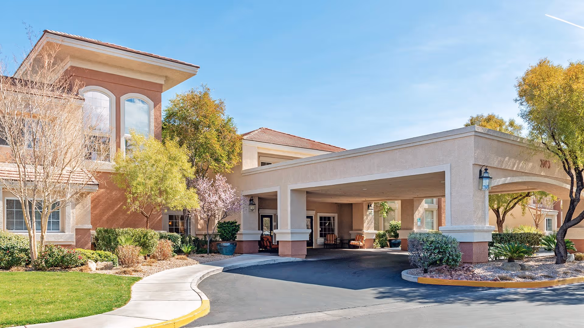 Exterior view of Atria Seville senior living facility showing the entrance with a covered drop-off area, surrounded by landscaped greenery and trees under a clear blue sky.