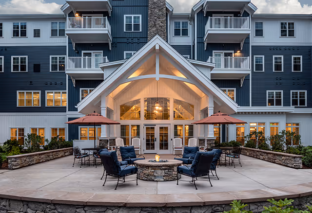 Exterior view of a senior living facility building at dusk with a covered entrance, multiple balconies, and large windows. In front of the entrance is a spacious patio area with cushioned chairs arranged around a stone fire pit and tables with umbrellas.