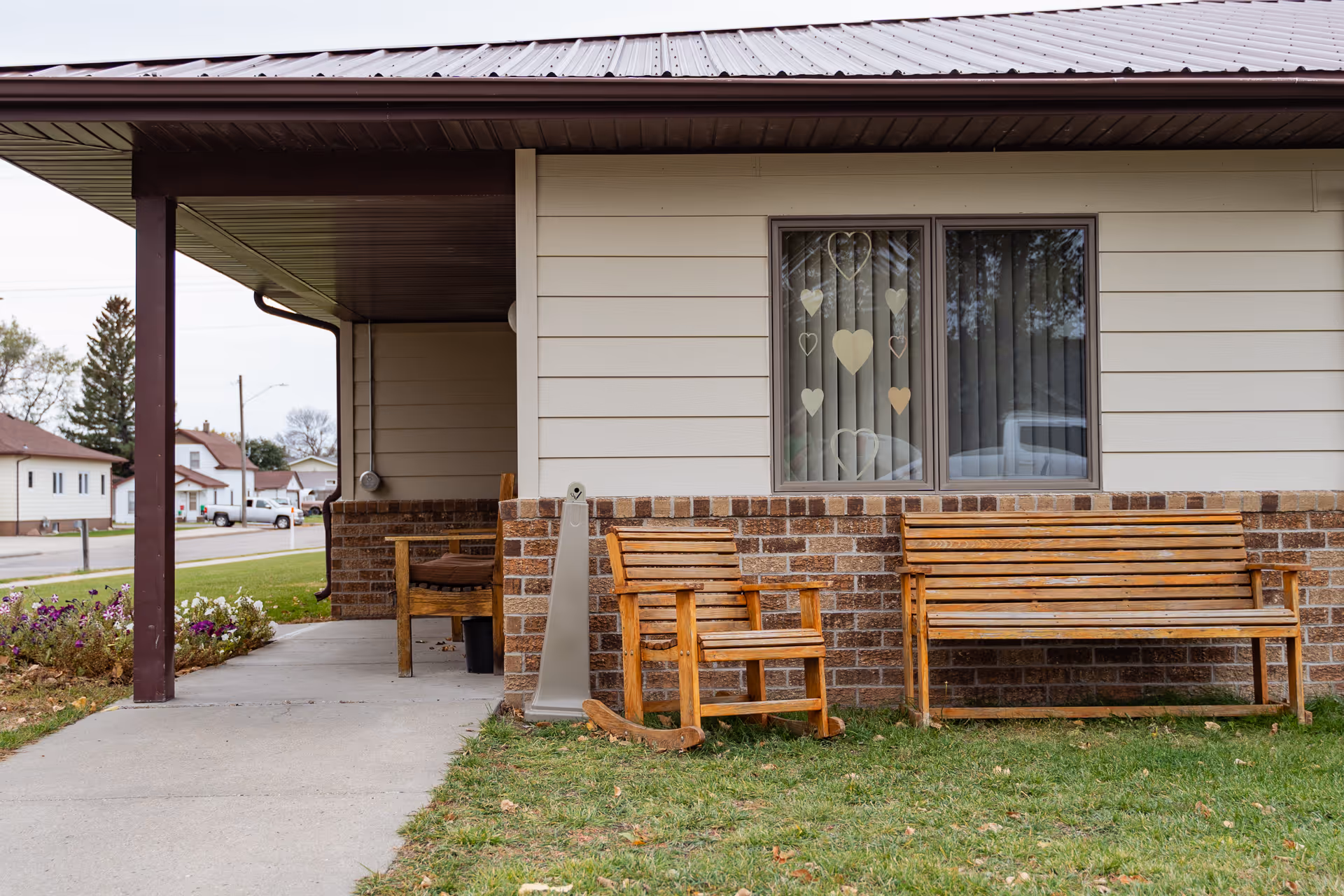 Single-story building exterior with a covered walkway, wooden benches and a rocking chair on the lawn in front of a window decorated with heart-shaped cutouts.