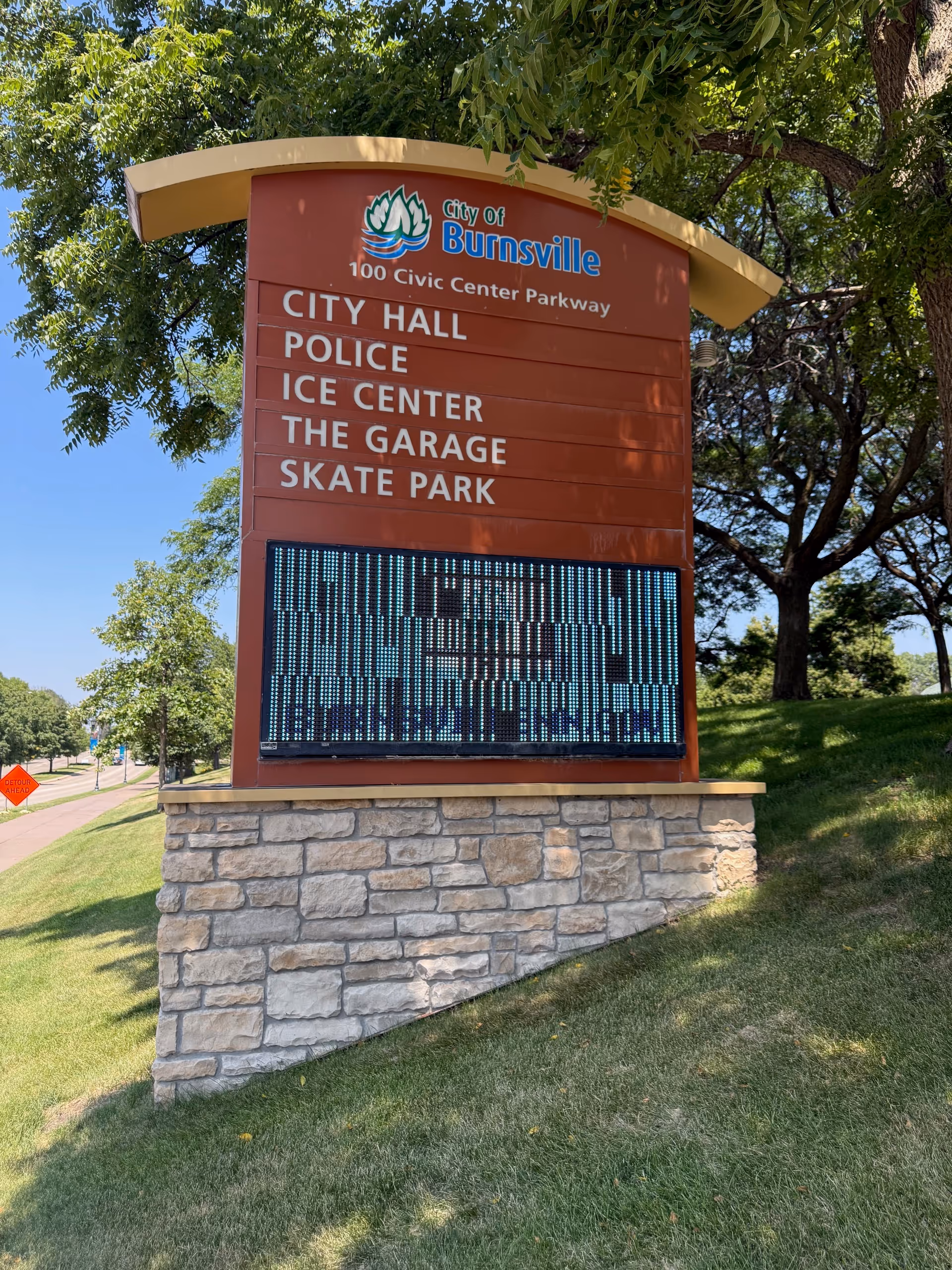 Outdoor city sign with listings and a digital display on a stone base.