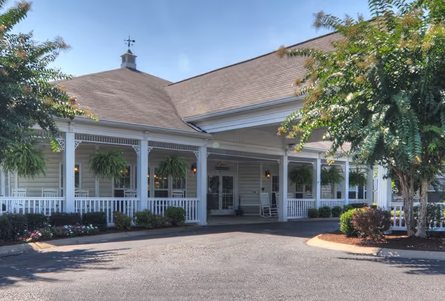 Front exterior view of a senior living facility with a covered porch, white railings, hanging plants, rocking chairs, and surrounding greenery under a clear blue sky.