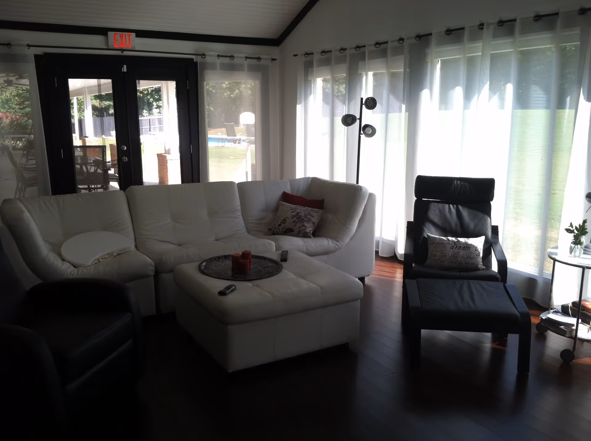 A living room with a white sectional sofa, a black armchair, and a black lounge chair with an ottoman. There is a white ottoman in the center with a tray holding red candles and a remote control. Large windows with sheer white curtains let in natural light, and a glass door leads outside to a patio area with outdoor furniture and a basketball hoop visible in the background.