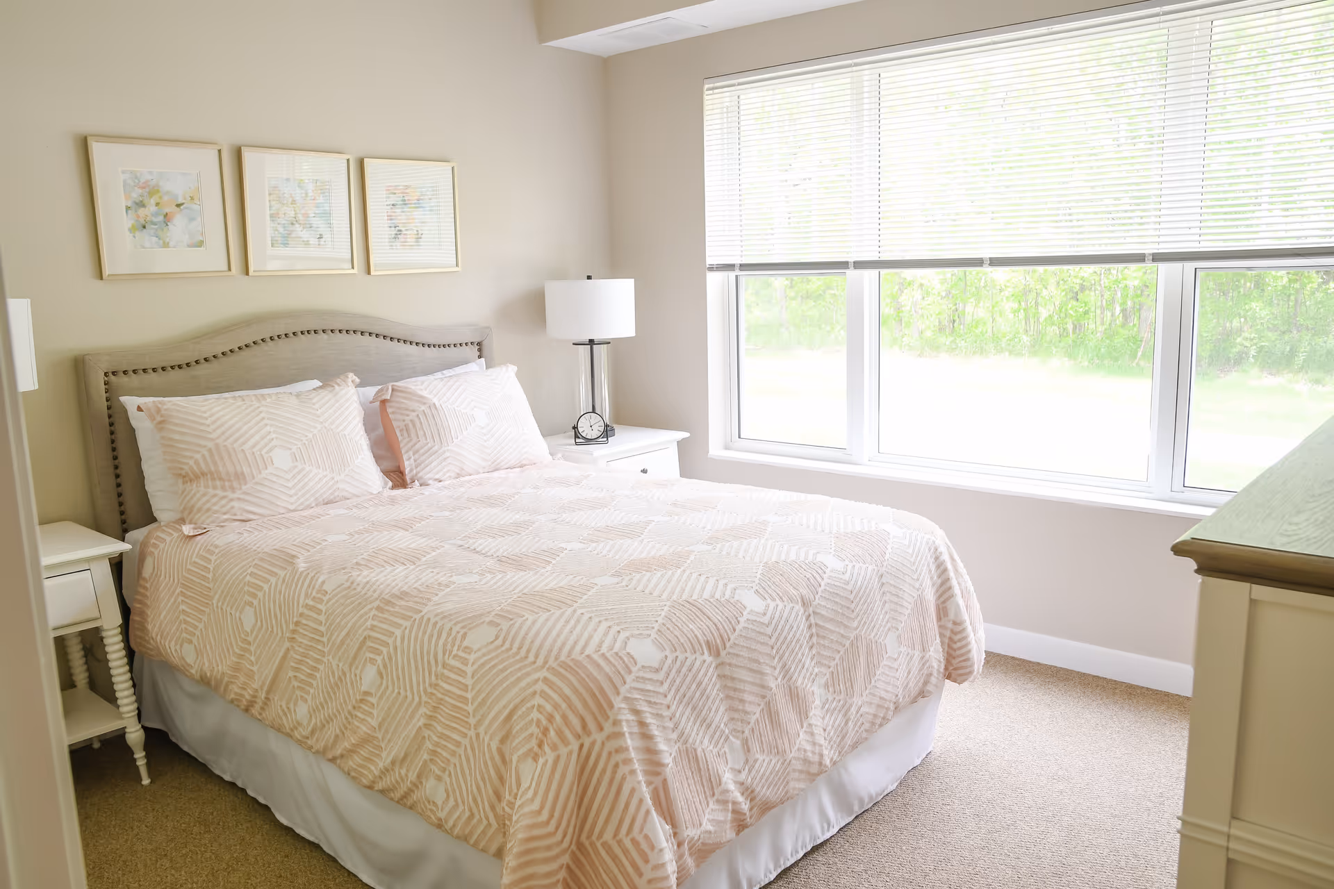 Bright bedroom with bed, two nightstands, lamp, and a large window overlooking greenery.