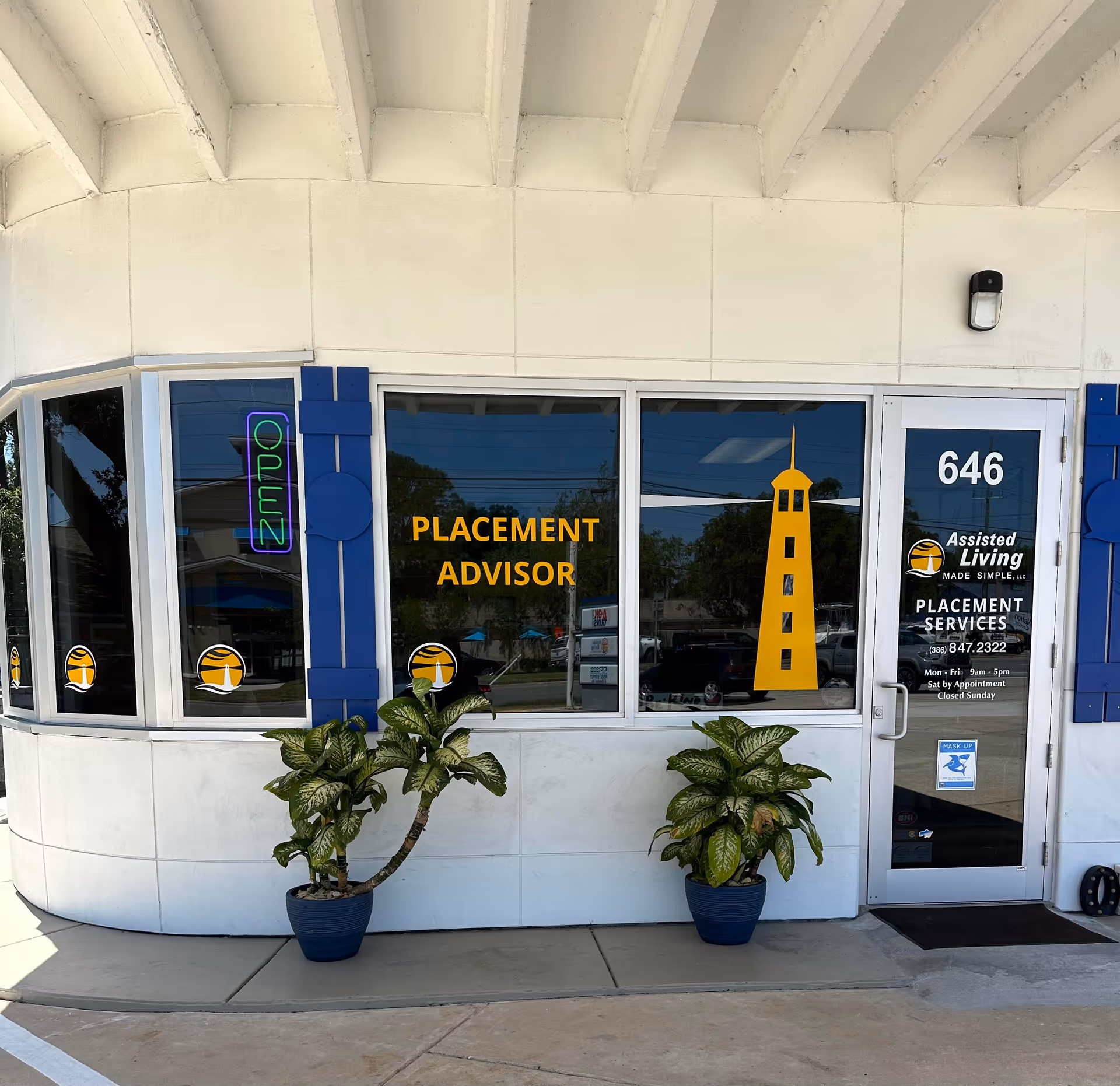 Exterior storefront with large windows, bright yellow placement advisor sign, and potted plants.