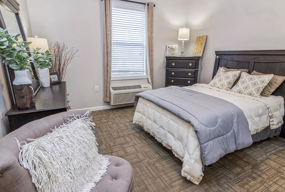 A neatly furnished bedroom with a bed, dresser and nightstand, an armchair with a decorative pillow, and a window with blinds.
