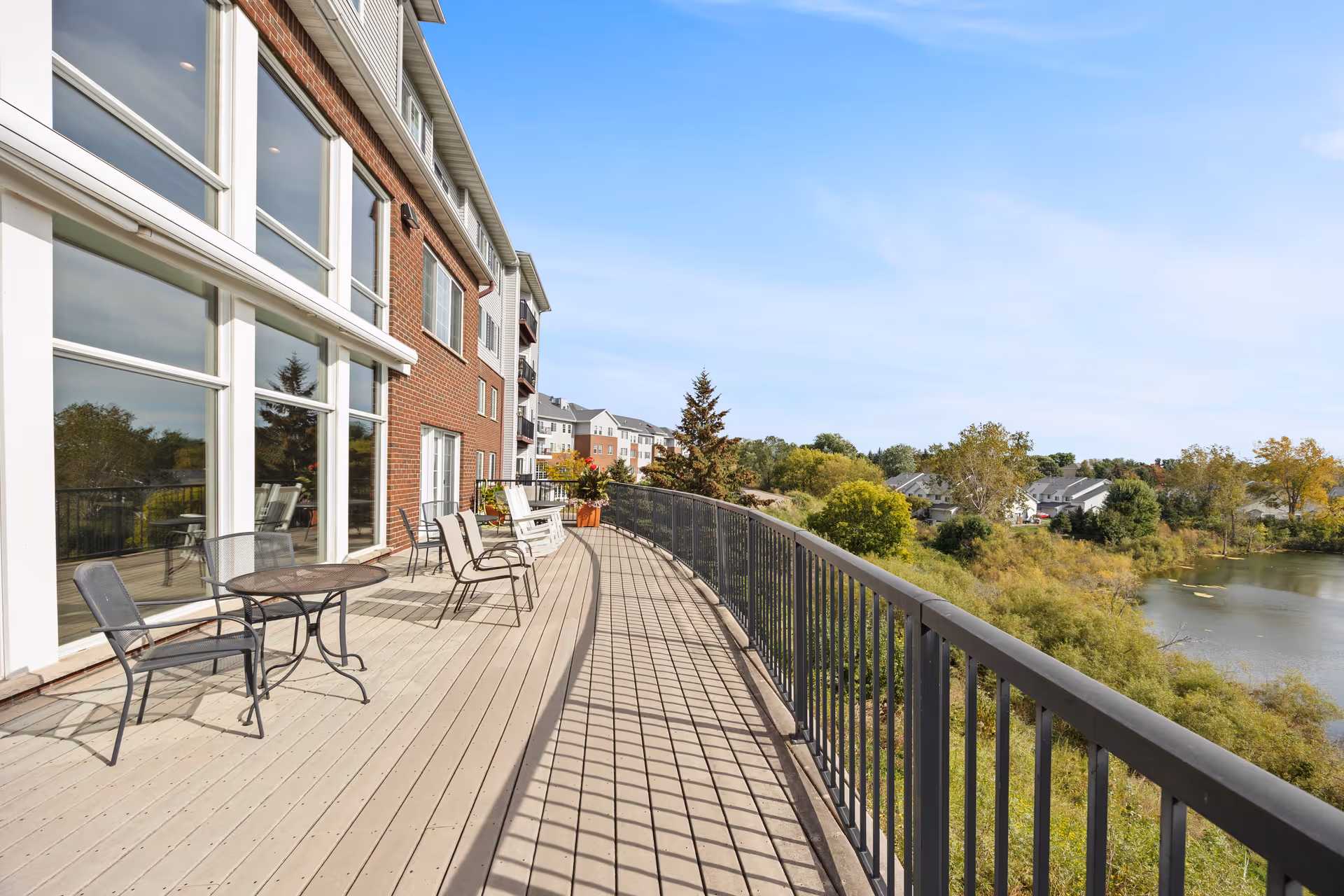 A spacious outdoor balcony with metal chairs and tables along the railing, attached to a multi-story brick building. The balcony overlooks a scenic view of trees, houses, and a body of water under a clear blue sky.